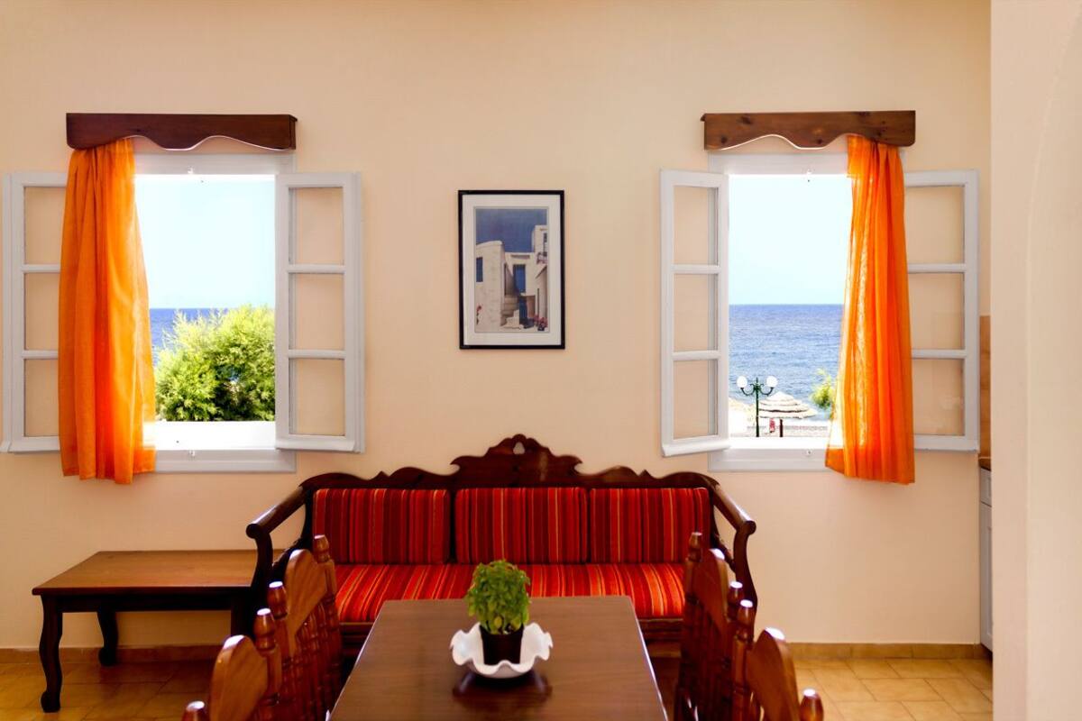 A dining room with a wooden table, red upholstered sofa, and two windows with orange curtains.