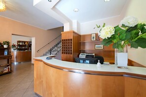 A reception area with a curved wooden reception desk, a staircase, a reception desk with a computer and printer, and a large white flower vase. - Hotel Stazione (Livorno)
