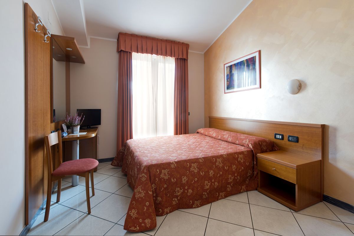 A hotel room with a bed, a desk with a chair, a TV, and a painting on the wall.