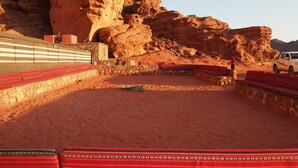 A desert setting with a large rock formation, a paved area with red benches, and a vehicle parked nearby. - Wadi Rum Candles Camp (Wadi Rum)