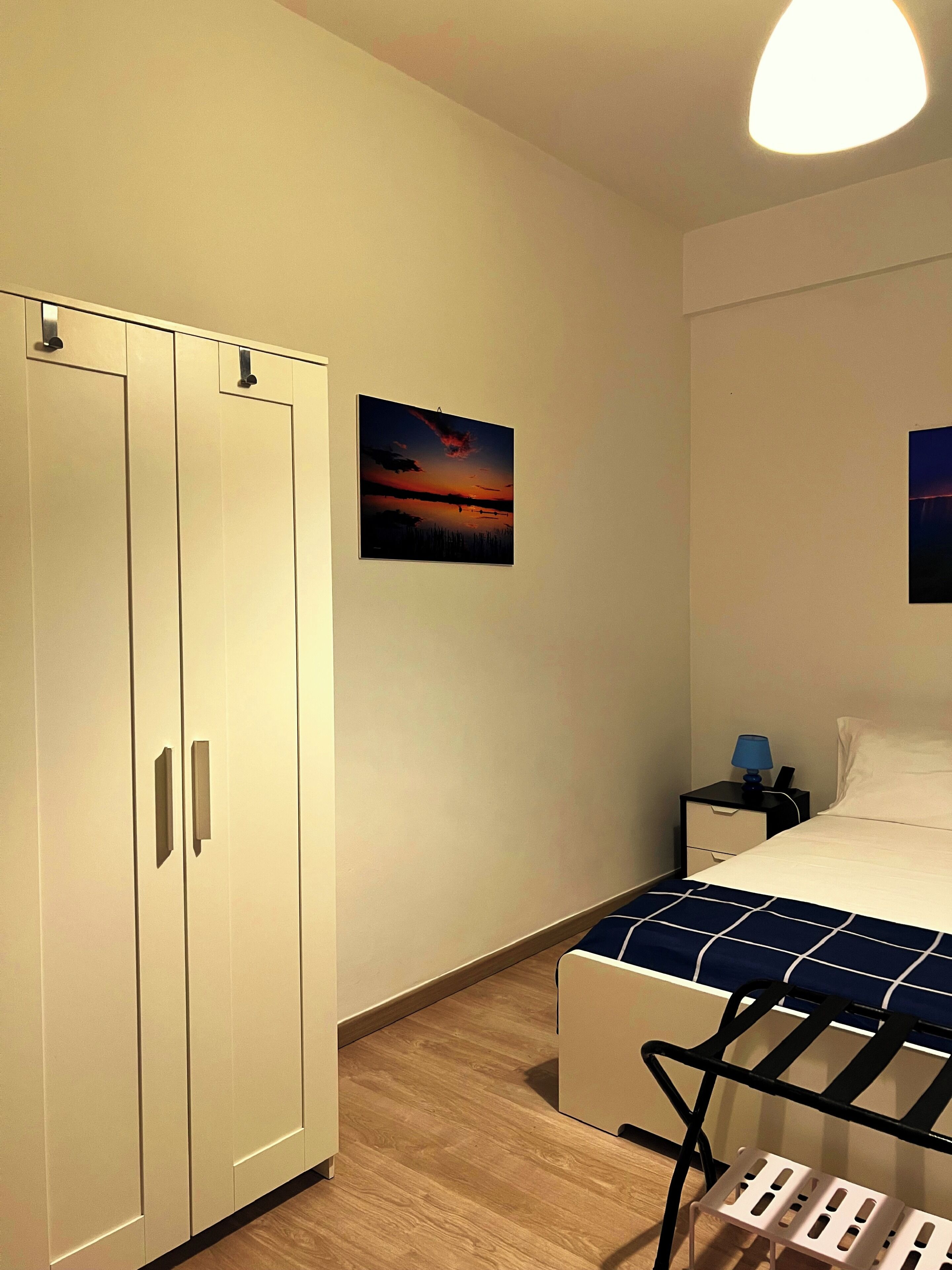 Een slaapkamer met een bed, nachtkastje, lamp, foto van een zonsondergang en een kledingkast.