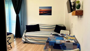 A room with a bed, a table with a tablecloth, a TV, a wall-mounted shelf with a plant, and a picture of a sunset. - B&B MaryJosè (Trapani)