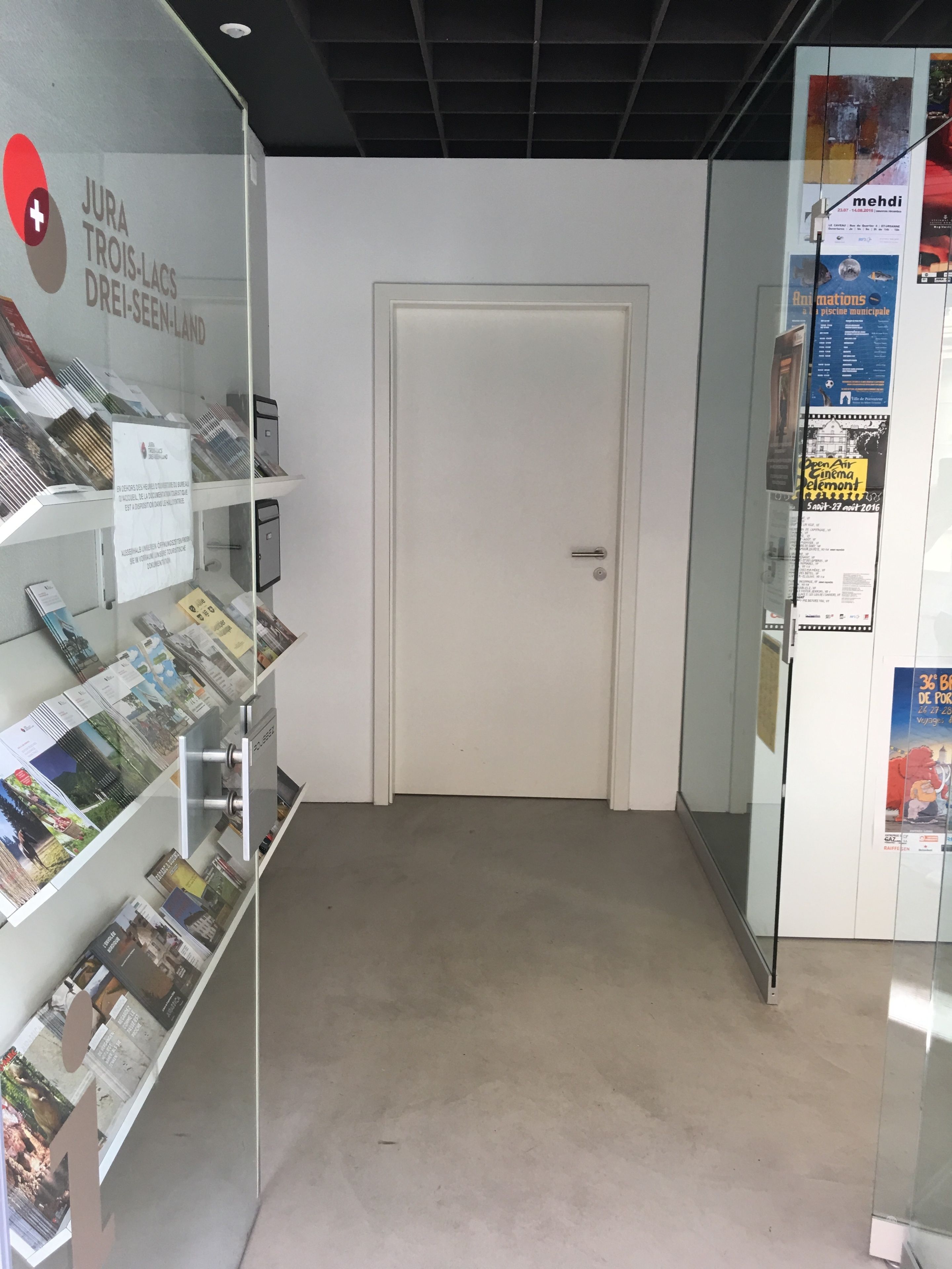 Un couloir avec des vitrines contenant des brochures, une porte fermée et des affiches sur le mur.