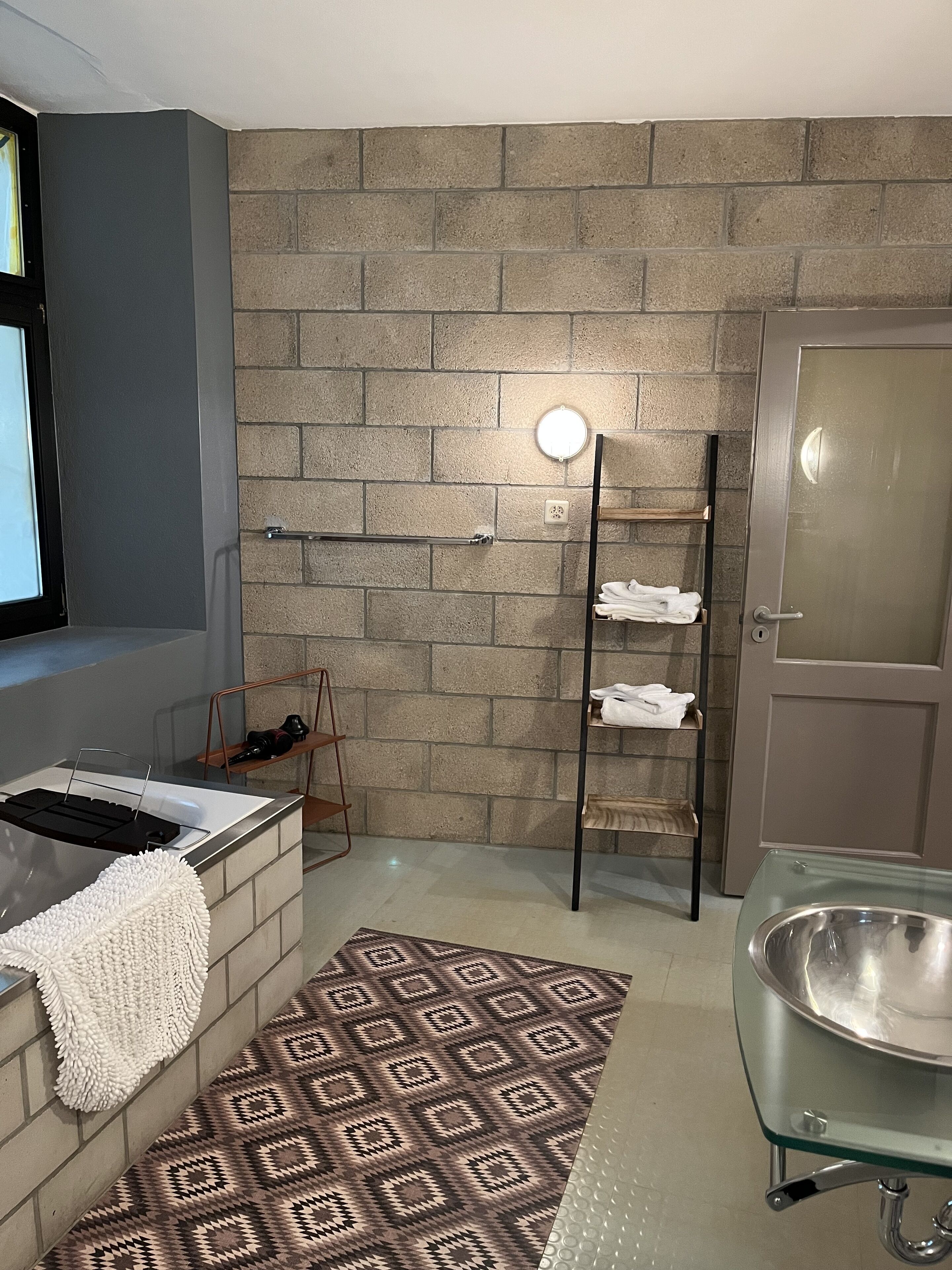 A bathroom with a bathtub, a towel rack, a patterned rug, and a brick wall.