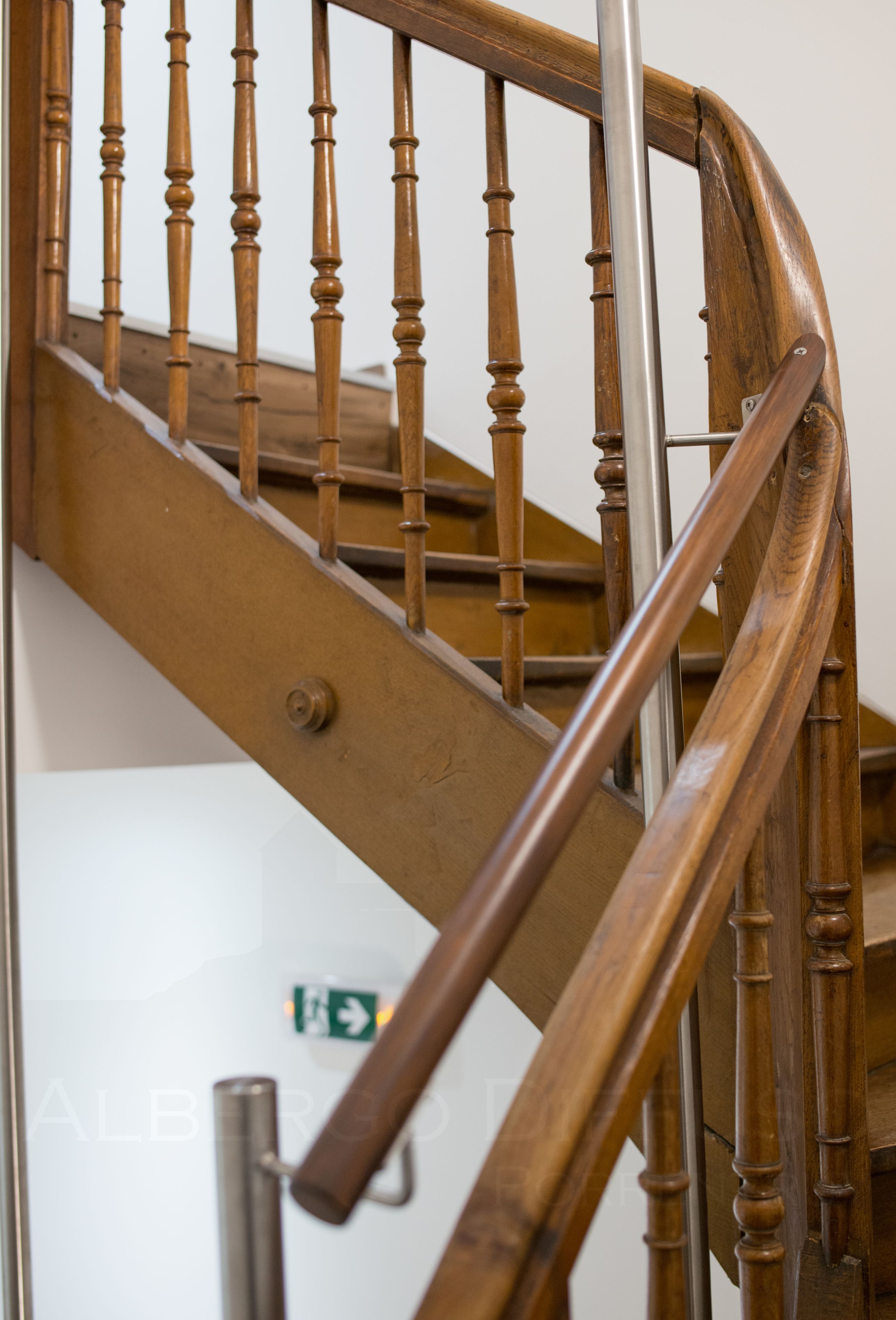 Una escalera de madera con barandilla de metal y una pared blanca con un letrero de salida de emergencia verde.