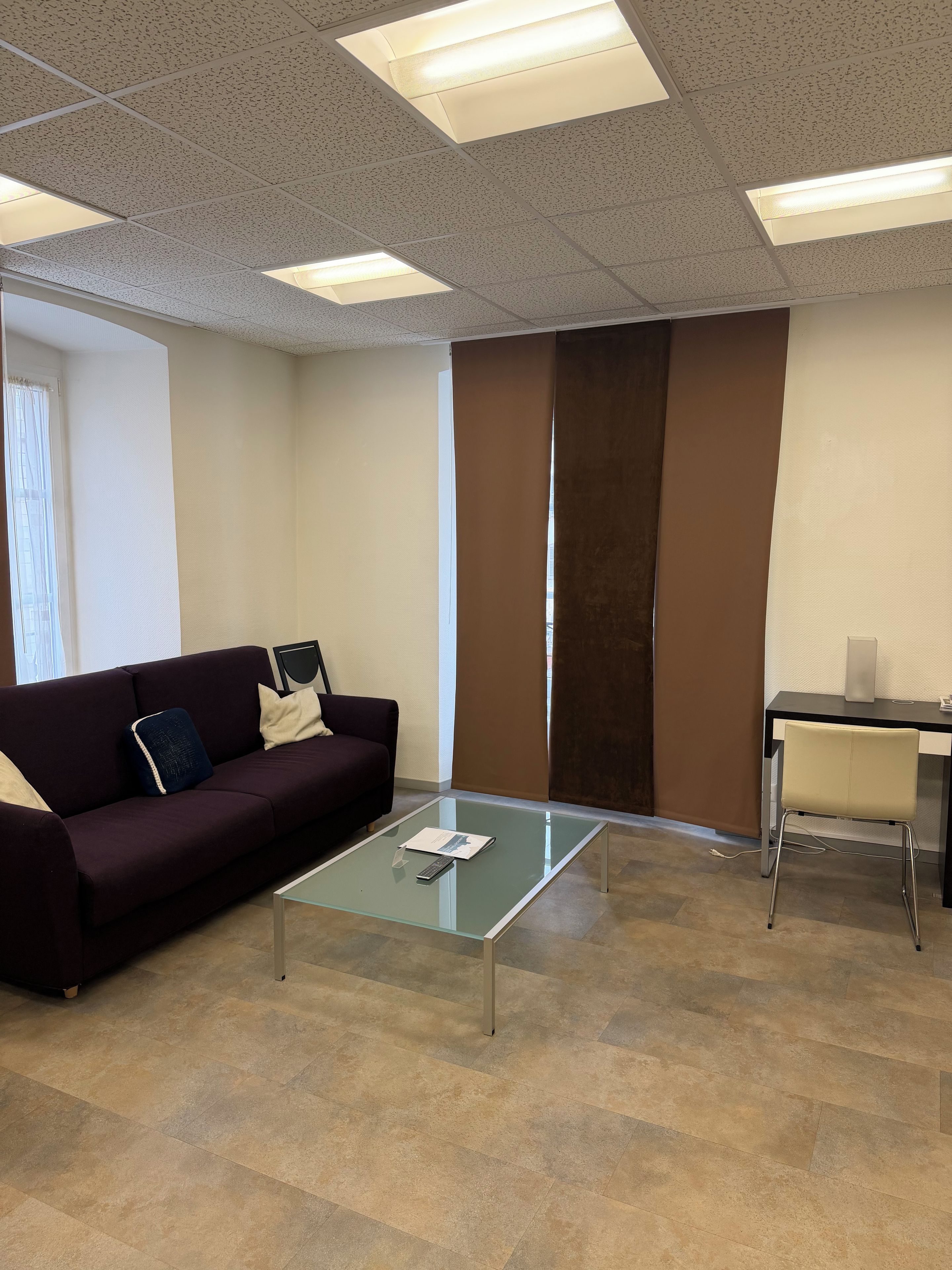 A modern office space with a glass-top table, a dark-colored sofa, and a desk with a chair.