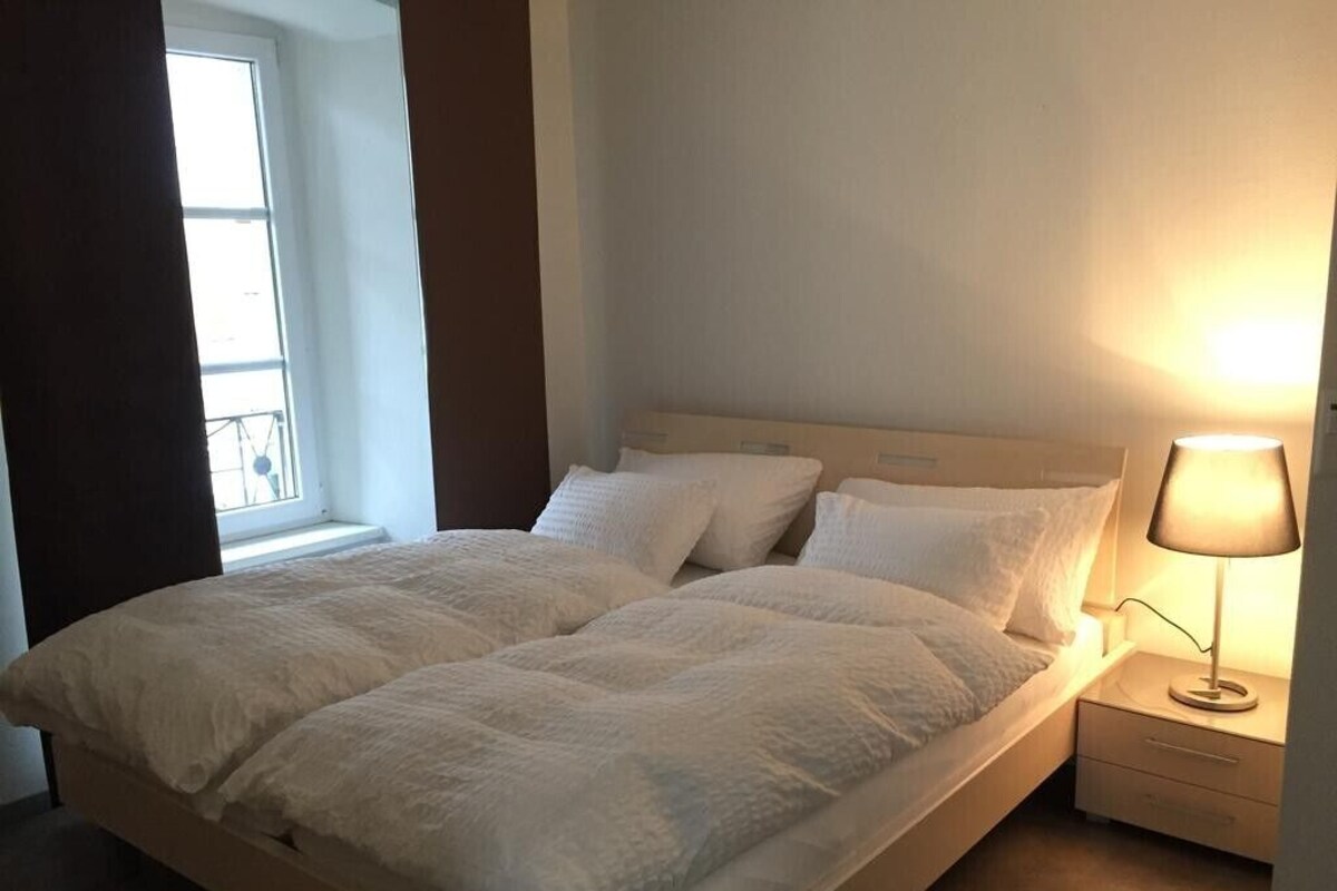 A neatly made double bed with a headboard, a bedside table with a lamp, and a window with curtains.