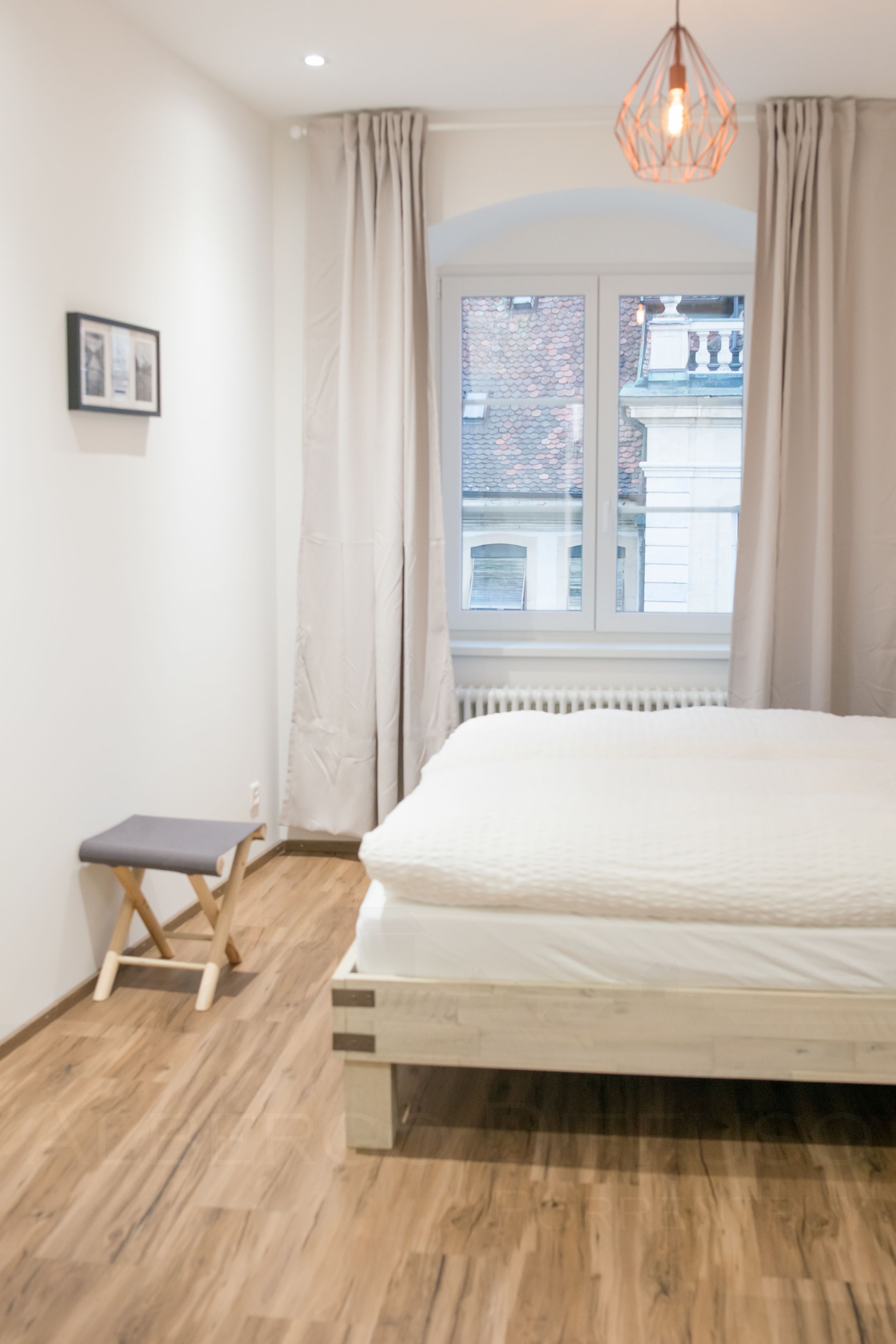 A bedroom with a wooden floor, a bed with a white mattress, a small bench, a window with curtains, and a hanging light fixture.