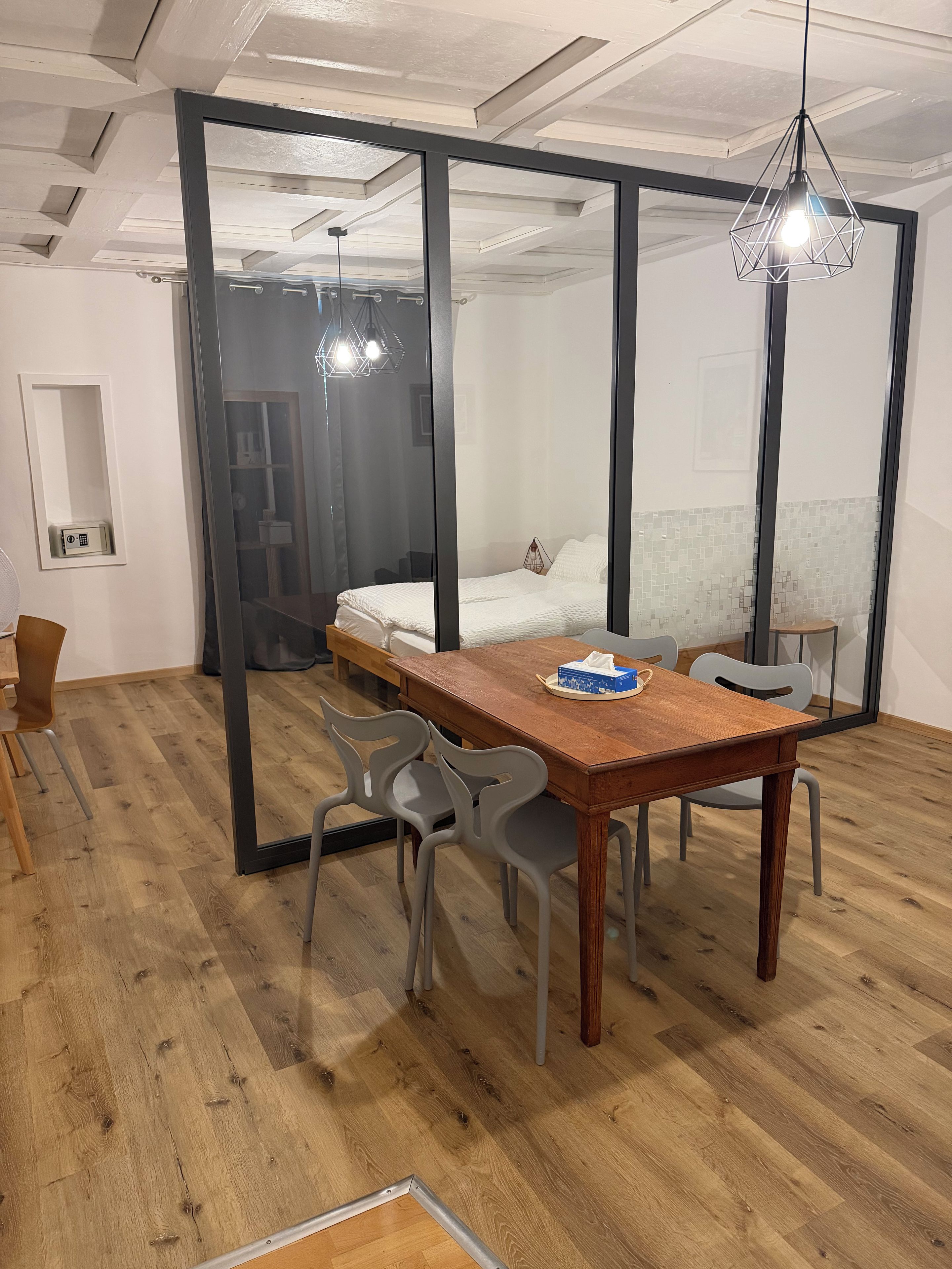 Una habitación moderna con una mesa de comedor de madera, sillas grises, una cama con ropa blanca y un separador de vidrio.