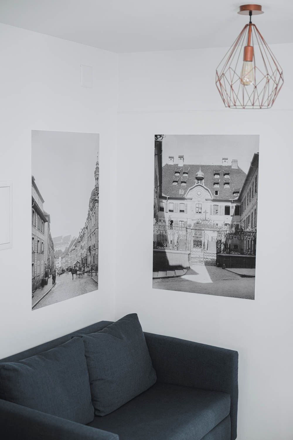 A room with a dark-colored sofa, two black and white photographs of buildings, and a geometric pendant light.
