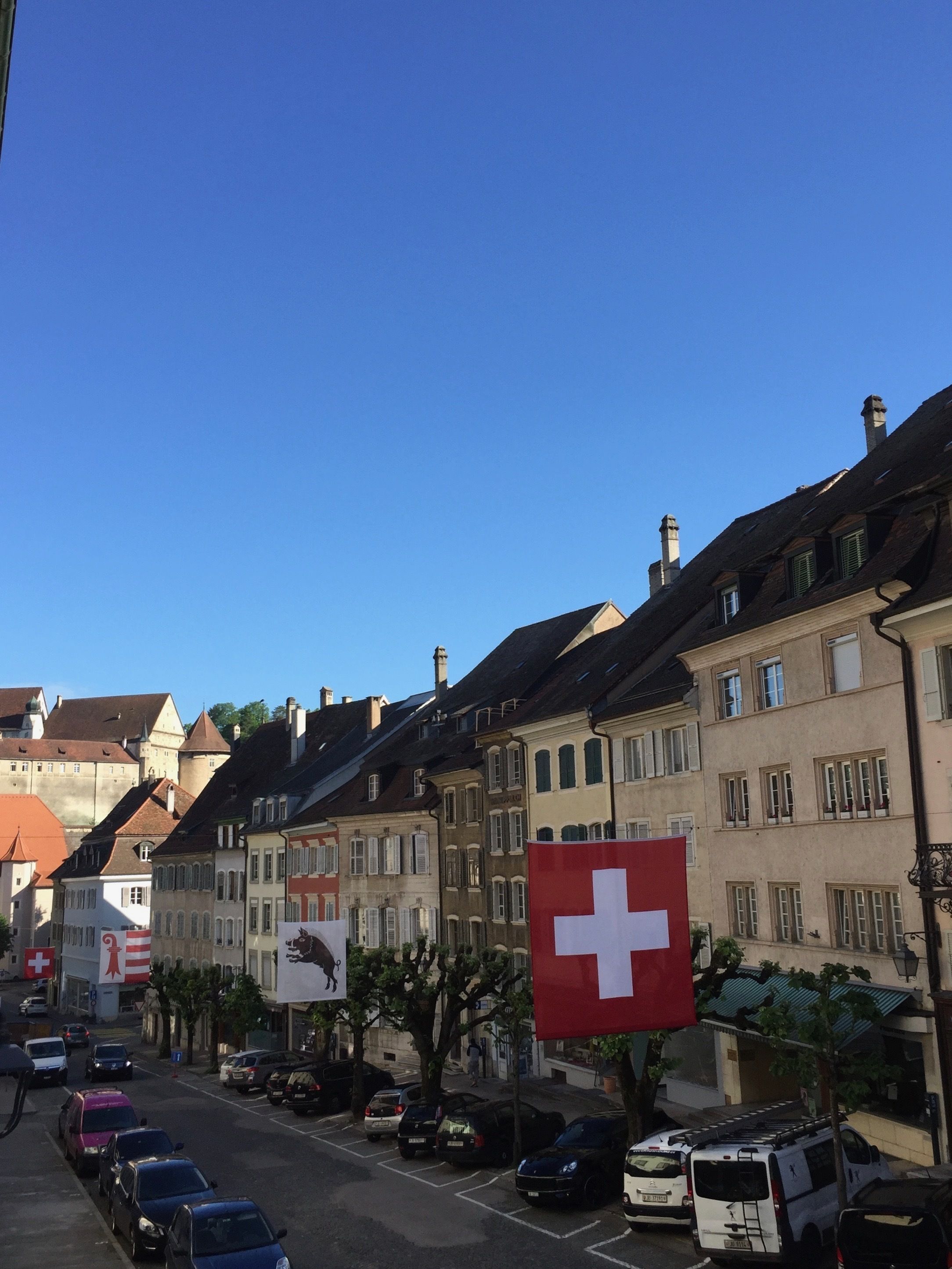 Une vue de rue avec des bâtiments européens traditionnels, des voitures garées et un ciel bleu dégagé.