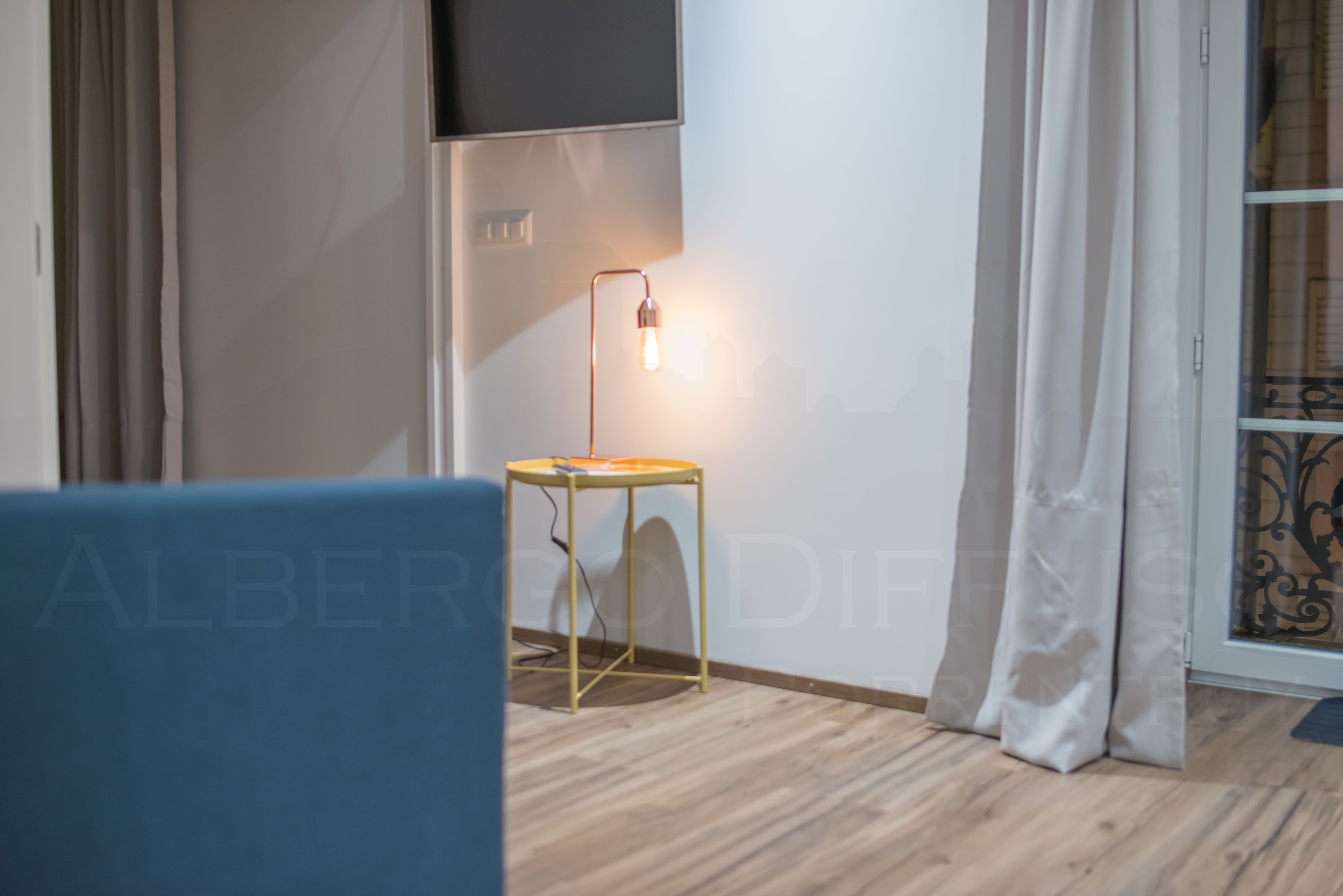 A room with a wooden floor, a small round table with a lamp, a blue chair, and a TV mounted on the wall.