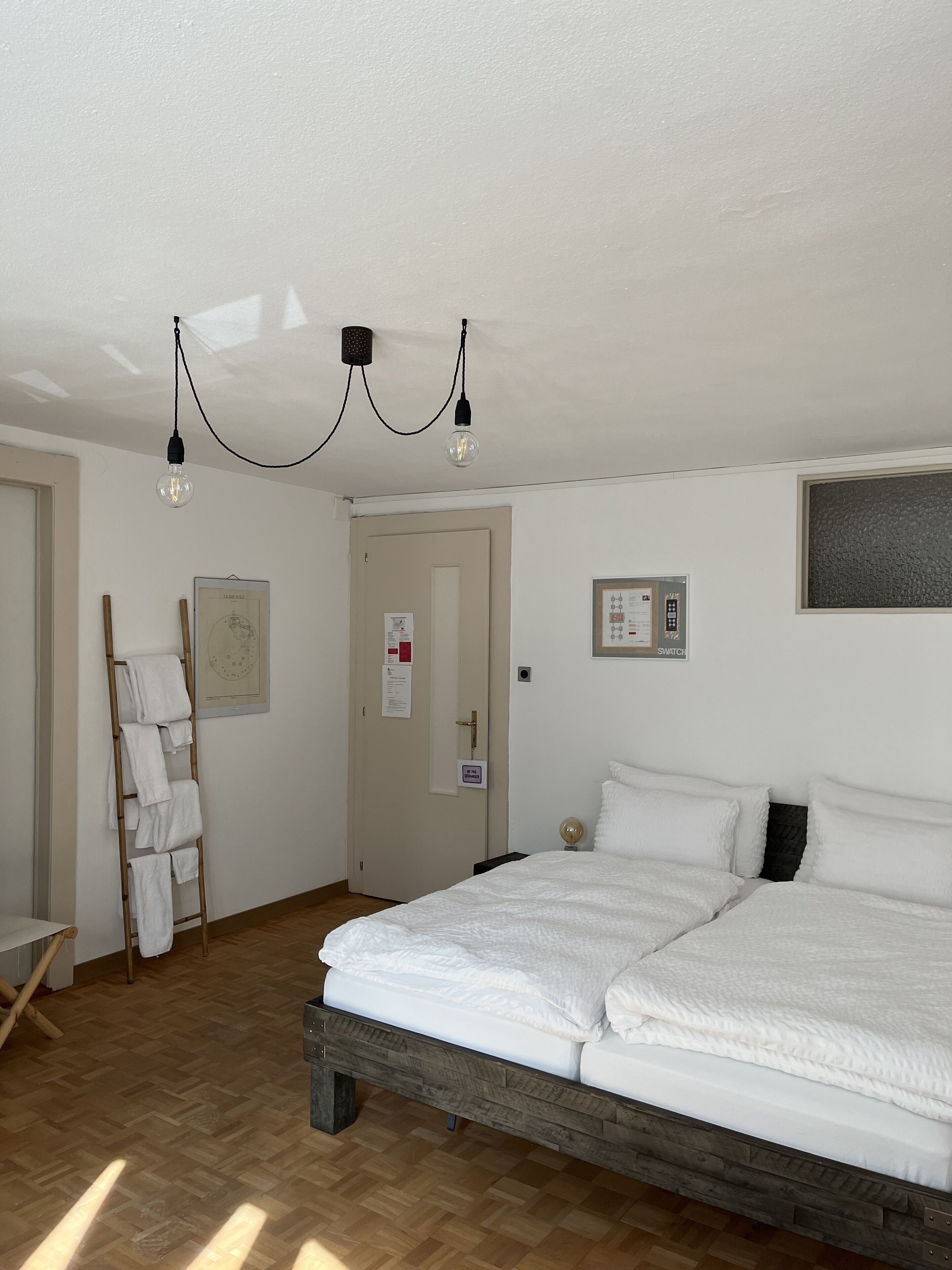 A bedroom with a wooden bed, white bedding, a ladder with towels, a door with a sign, and a light fixture.