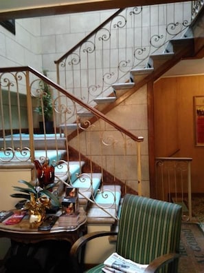 A staircase with ornate metal railings and a wooden handrail, leading to a landing with a green striped chair and a small table with decorative items. - Albergo Al Gobbo   (Venice)