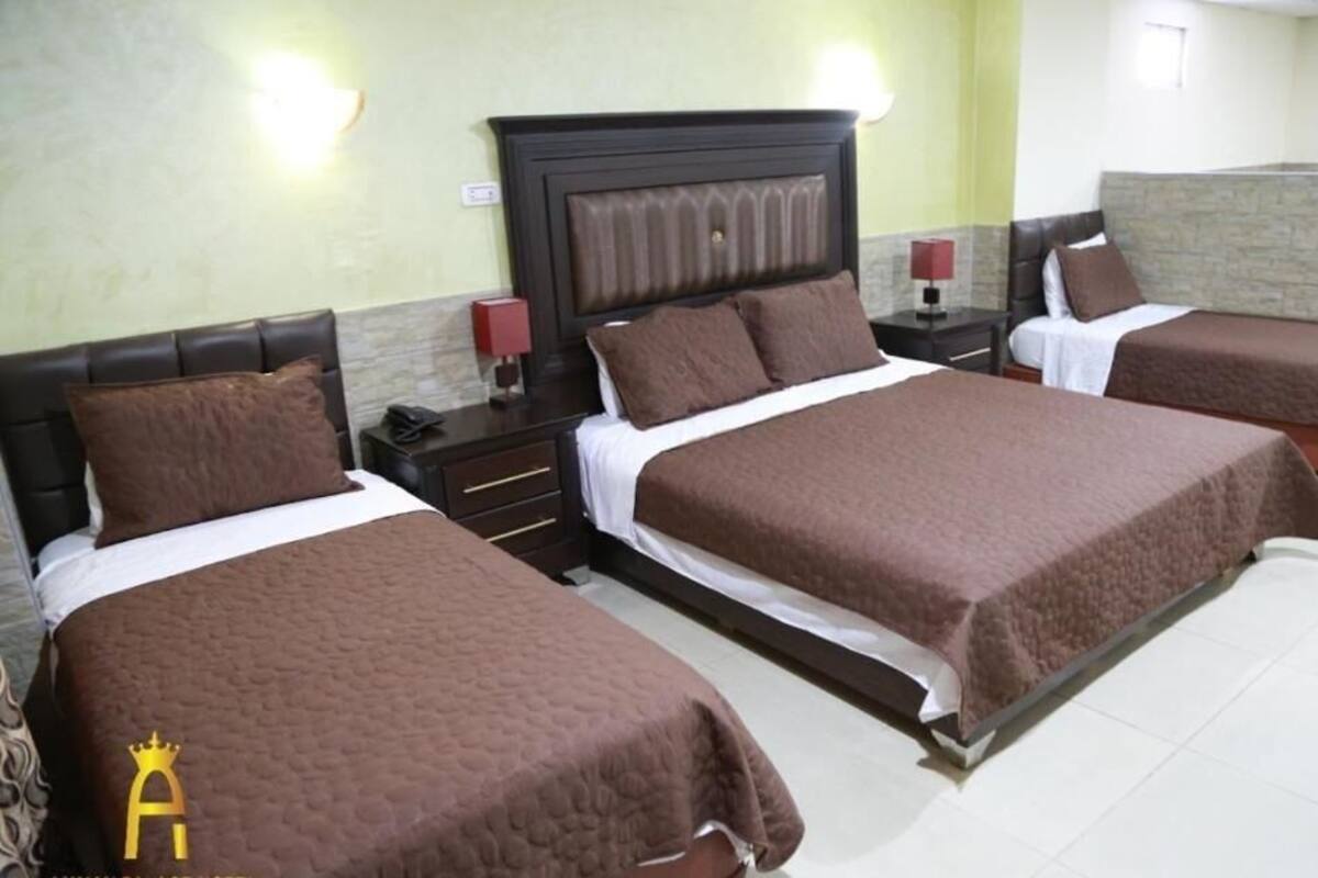 Une chambre d’hôtel avec trois lits, chacun recouvert d’une couverture marron et doté d’une tête de lit. On y trouve deux tables de chevet avec des lampes, et un mur en pierre décoratif.