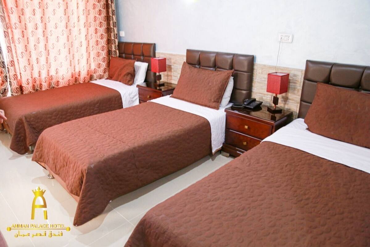 Une chambre d’hôtel avec trois lits, chacun recouvert d’une couverture marron et doté de draps blancs. On y trouve des tables de chevet en bois avec des lampes rouges, et un rideau à motifs est visible.