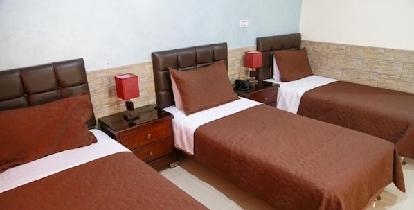 A hotel room with three single beds, each with a brown bedspread and matching pillows. There are bedside tables with lamps and a small drawer. The room has a tiled floor and a stone accent wall.