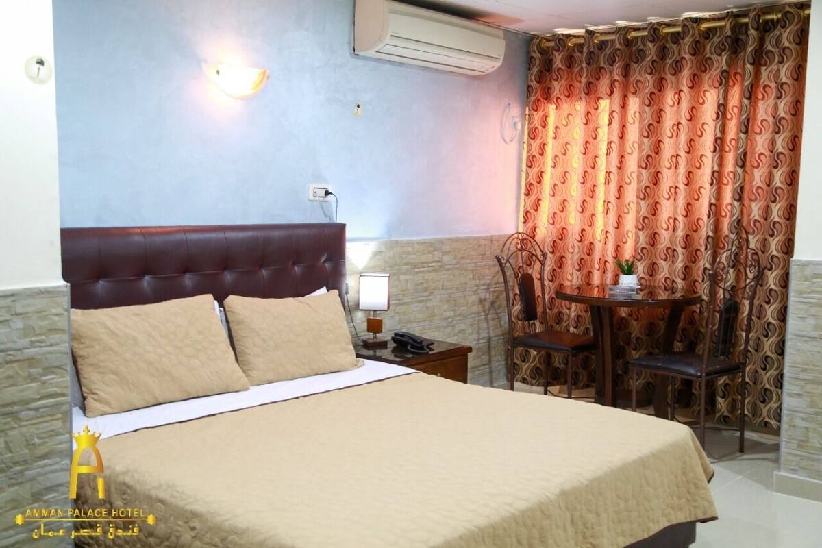 Une chambre d’hôtel avec un grand lit, une table de chevet avec une lampe, une chaise, une petite table avec une plante et un rideau à motifs.