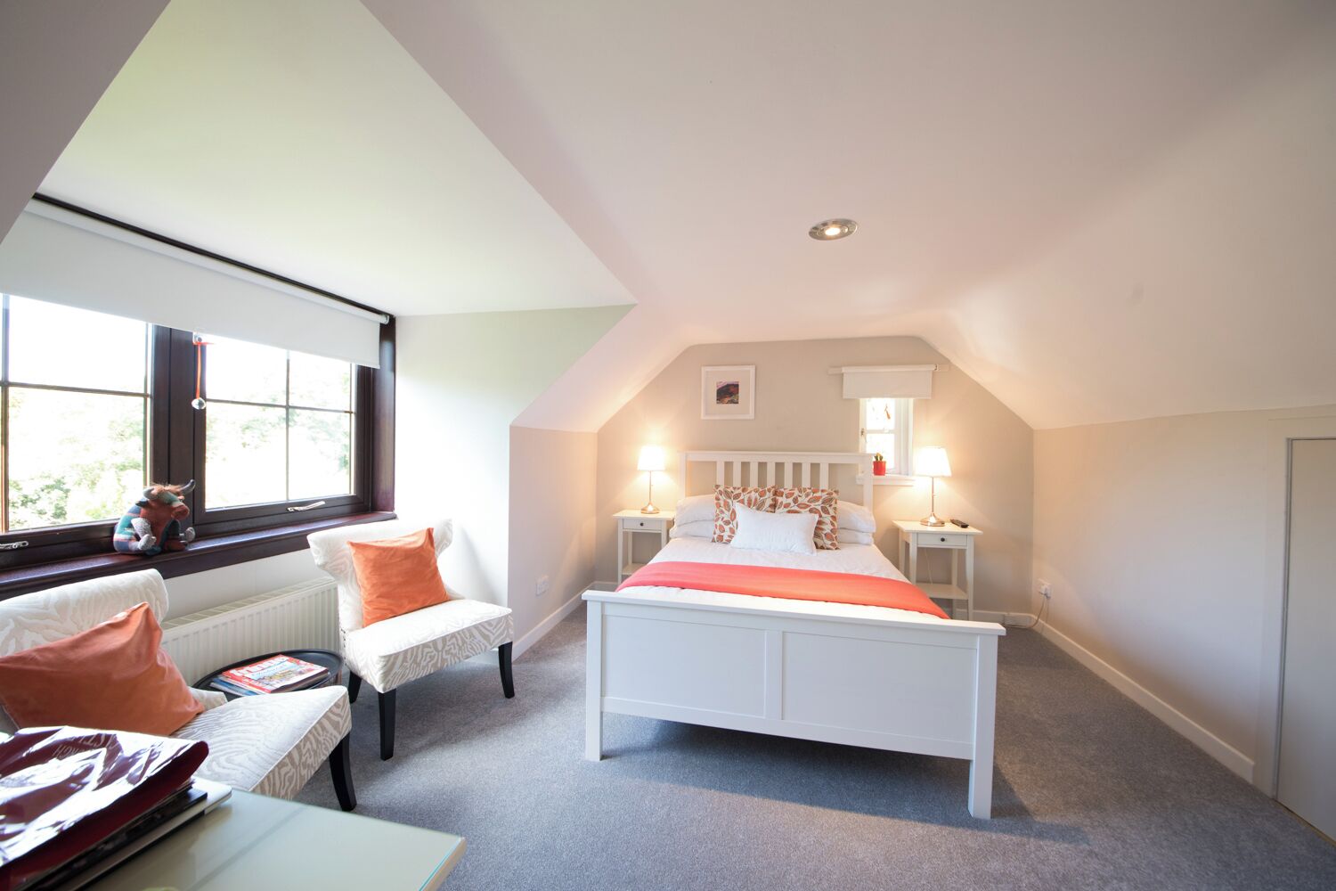 A bedroom with a sloped ceiling, a bed with white bedding, two bedside lamps, a window with a view, and a chair with a cushion.