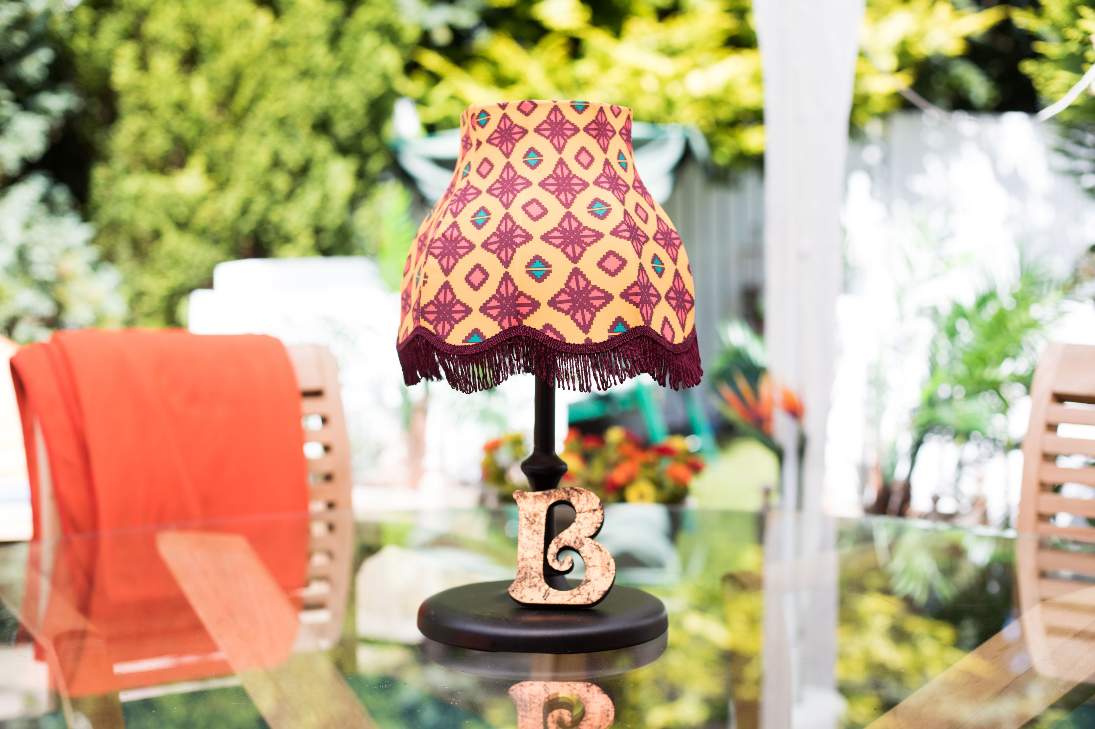 A table lamp with a patterned lampshade and a letter 'B' base, placed on a glass table with a blurred outdoor background.