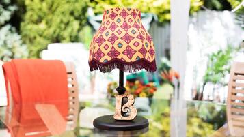 A table lamp with a patterned lampshade and a letter 'B' base, placed on a glass table with a blurred outdoor background.