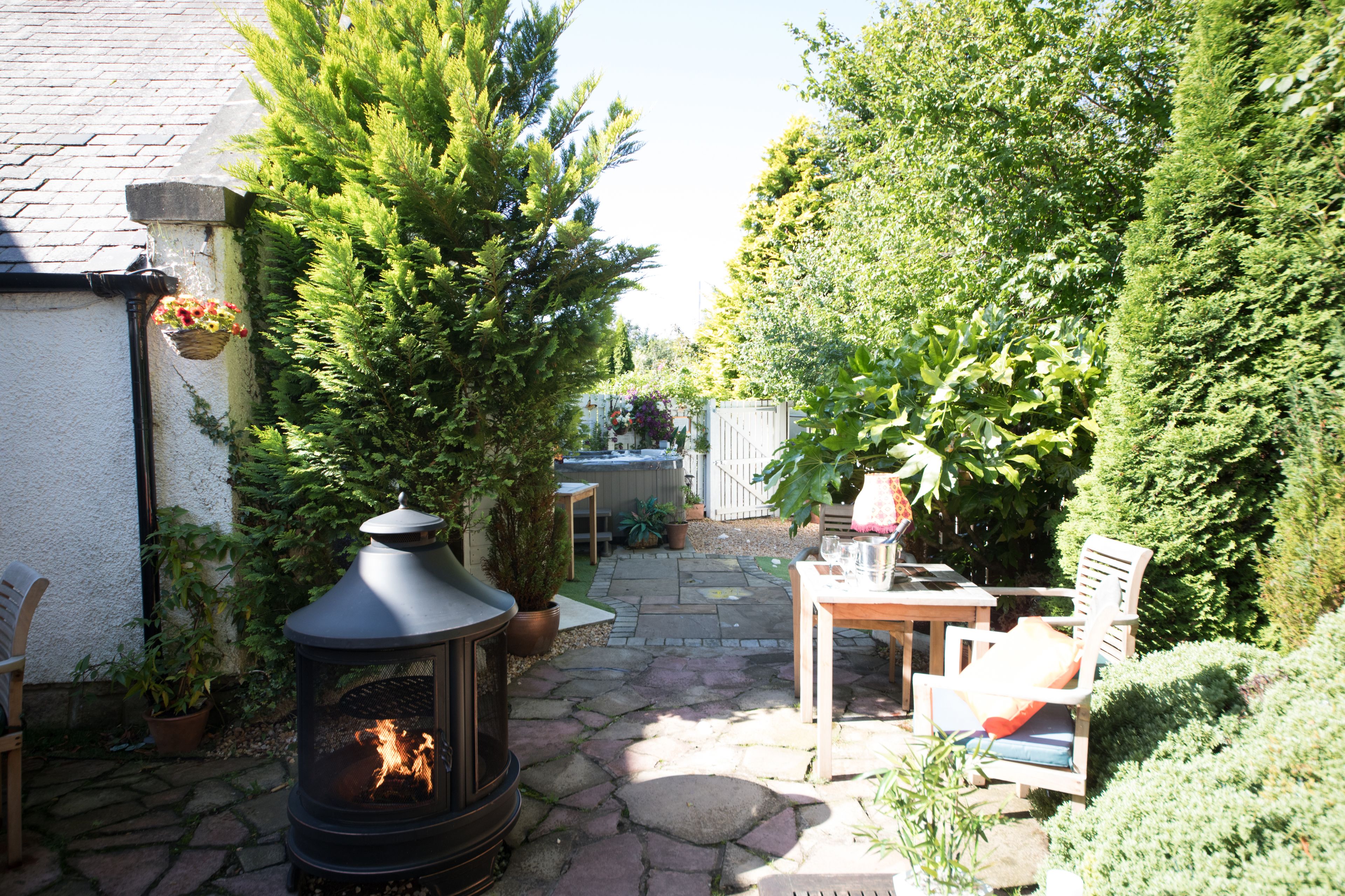 A stone-paved patio with a wood-burning stove, outdoor seating, and lush greenery.