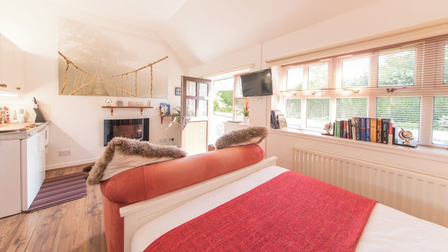 A bedroom with a bed, a TV, a window with blinds, a bookshelf, and a fireplace.