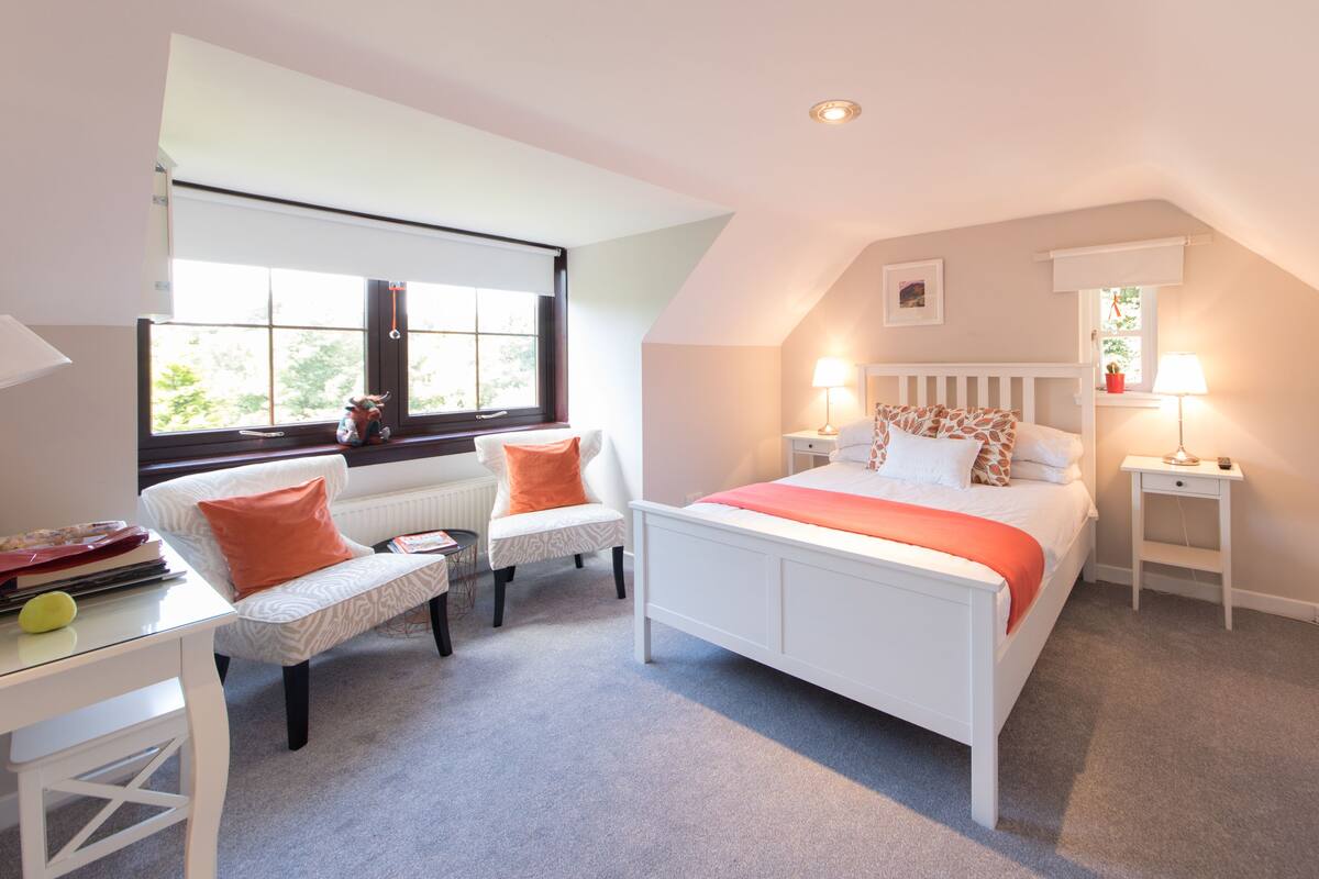 A bedroom with a white bed, two white chairs, a small table, and a window with blinds.