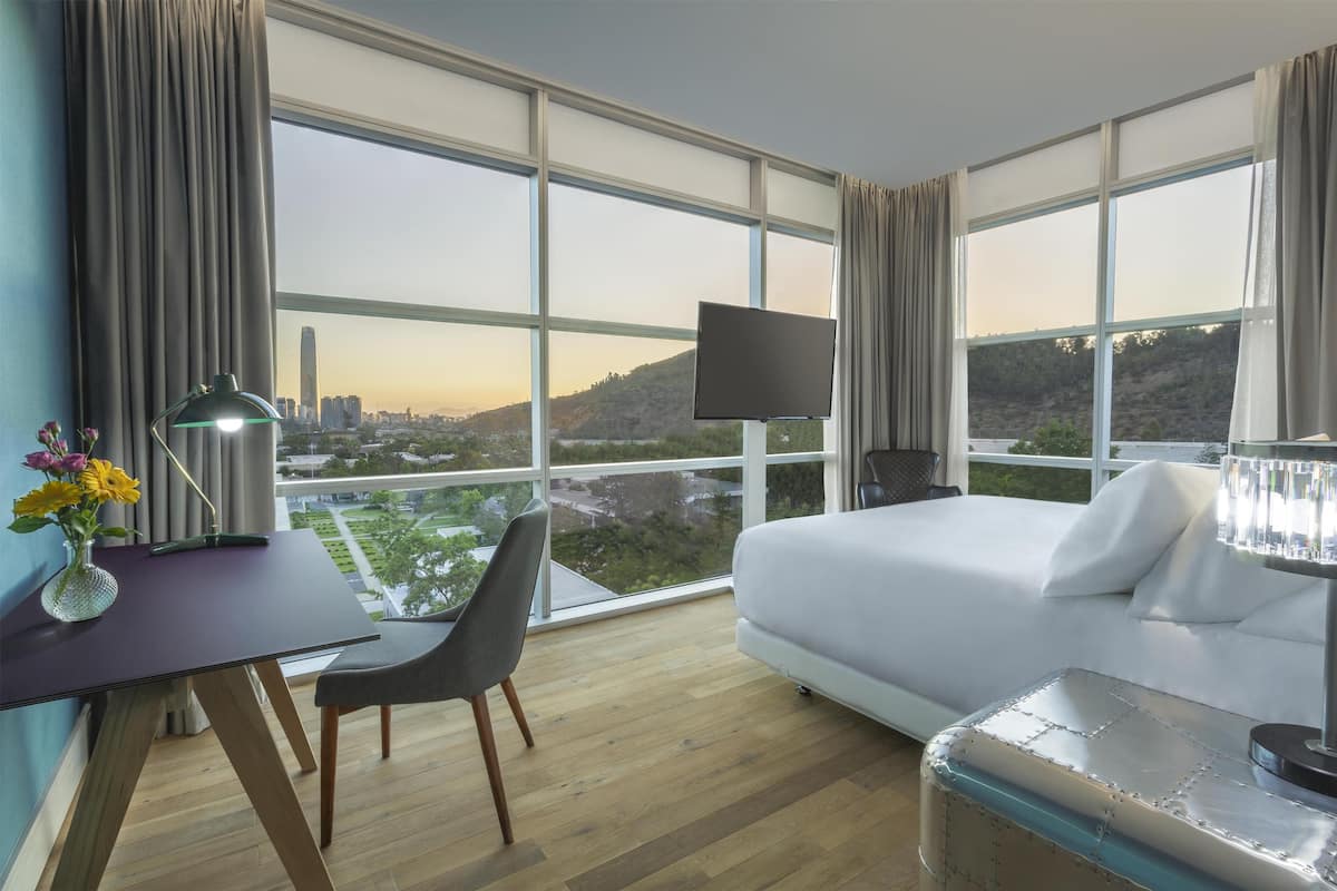 Ein Hotelzimmer mit großem Fenster, einem Bett, einem Schreibtisch mit Stuhl und einem Fernseher.