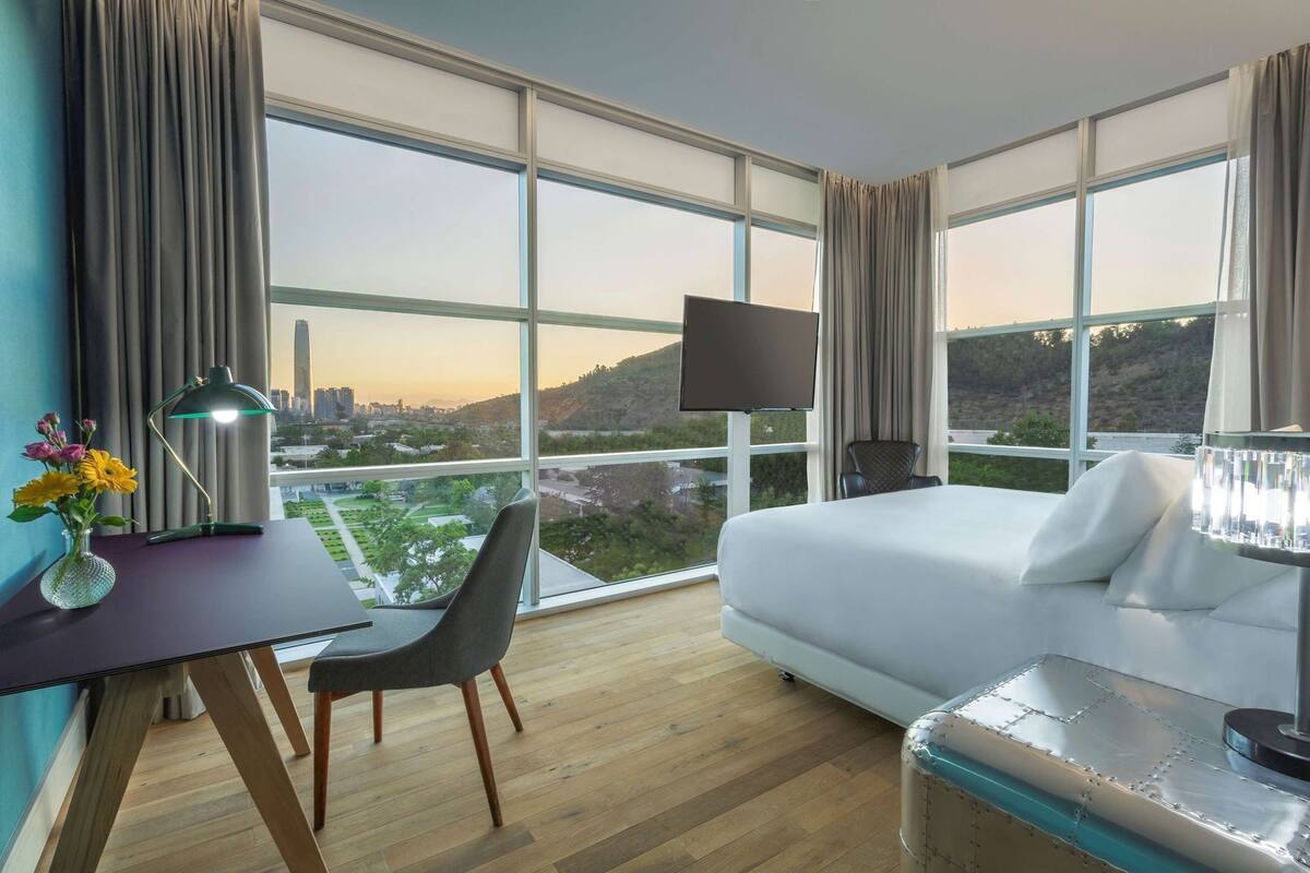 Ein Hotelzimmer mit einem großen Bett, einem Schreibtisch mit Stuhl, einem Fernseher und Blick auf die Stadt.