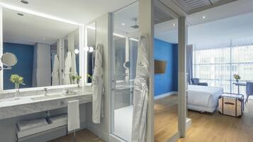 Un baño de hotel moderno con un espejo grande, cabina de ducha de vidrio y una cama en la habitación contigua.