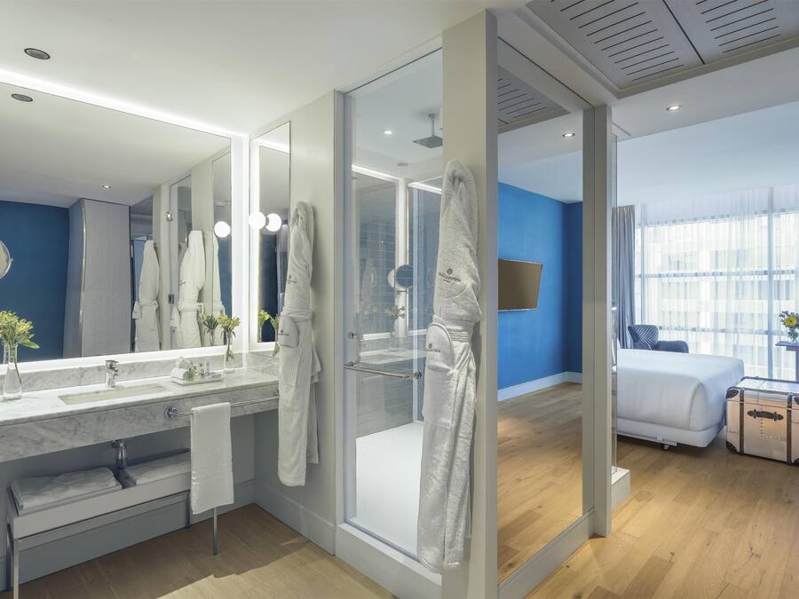 Un baño de hotel moderno con un espejo grande, cabina de ducha de vidrio y una cama en la habitación contigua.