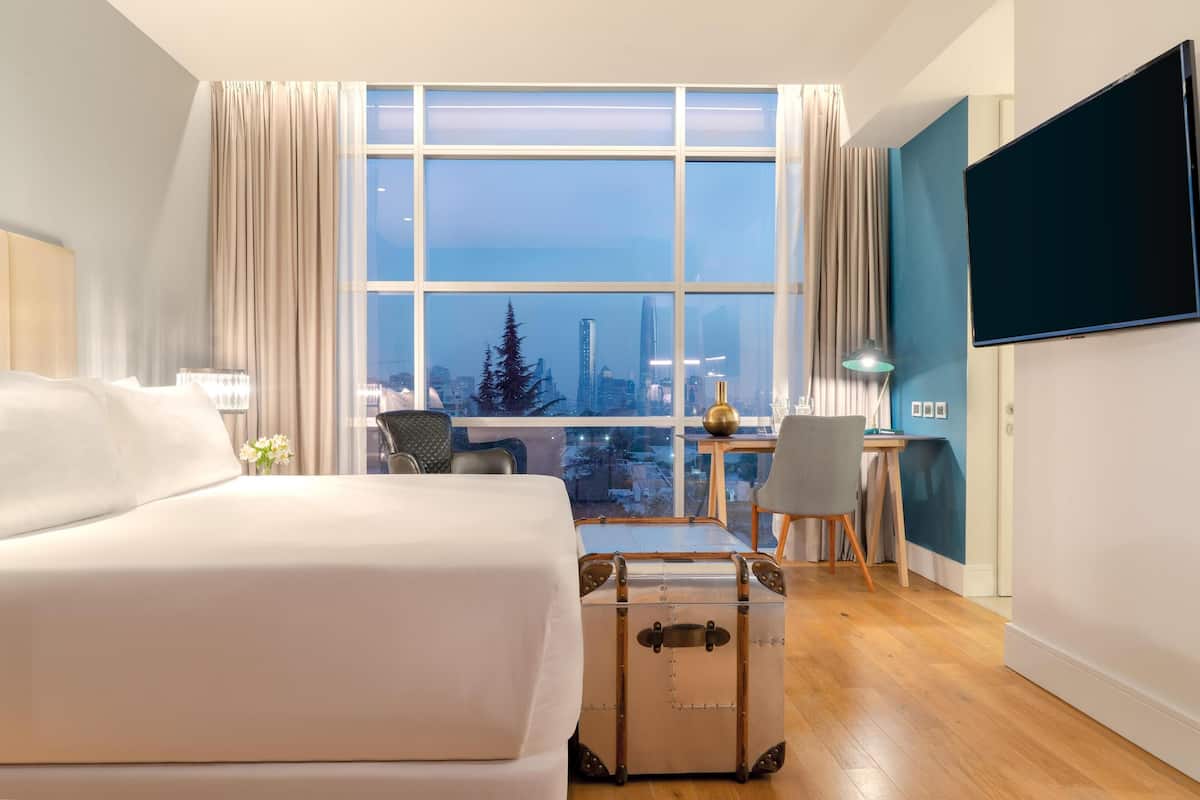 Ein Hotelzimmer mit einem großen Bett, einem Schreibtisch mit Stuhl, einem Koffer und einem an der Wand befestigten Fernseher.