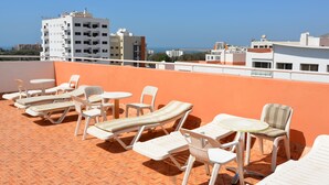A rooftop terrace with lounge chairs and tables, a clear view of the sea, and modern buildings in the background.