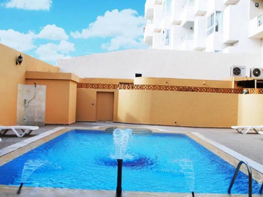 Una piscina con una fuente, rodeada de sillas de descanso y un edificio con balcones.