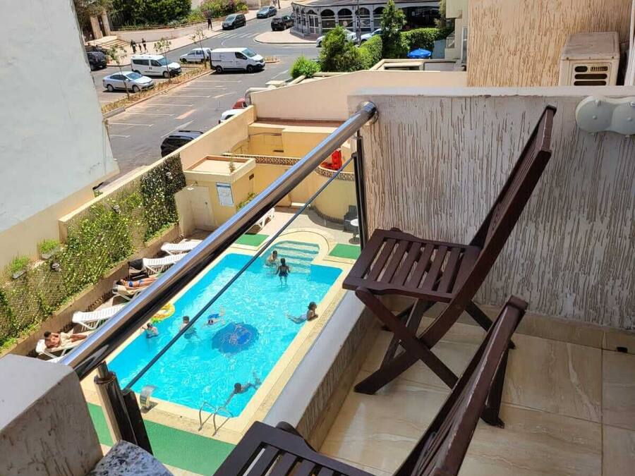 Un balcón con piscina, sillas de descanso y vistas a un paisaje urbano con edificios y automóviles estacionados.
