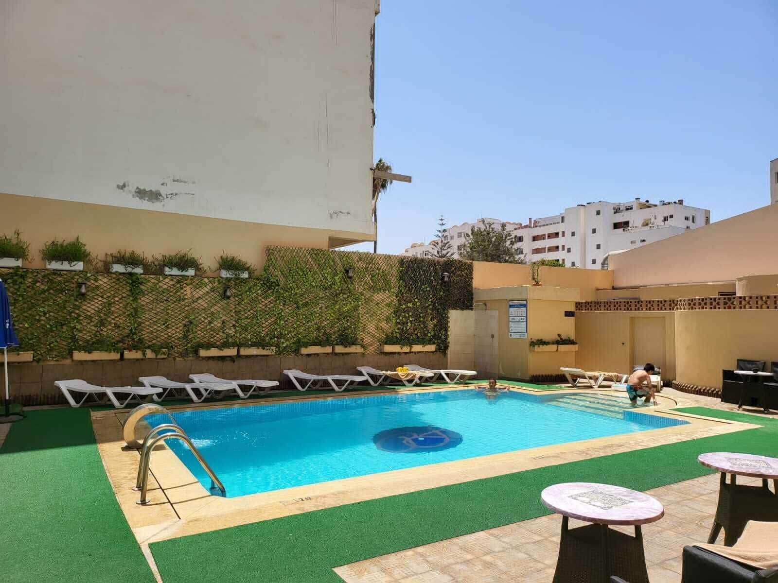 A swimming pool with lounge chairs and tables, surrounded by a low wall with greenery.