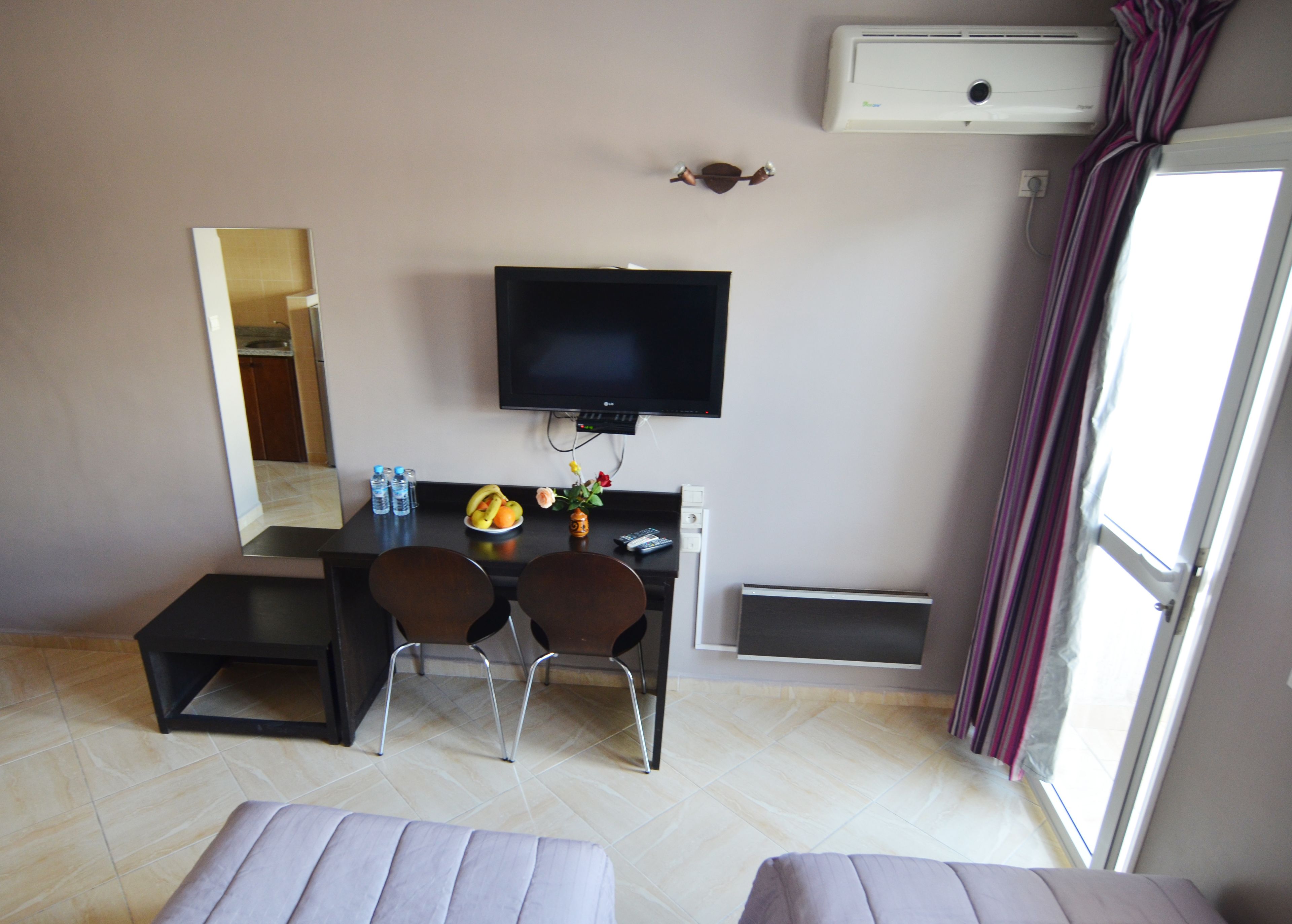 Una habitación de hotel con una cama, un escritorio con dos sillas, un televisor, una mesita con fruta y un florero, y una ventana con cortinas.