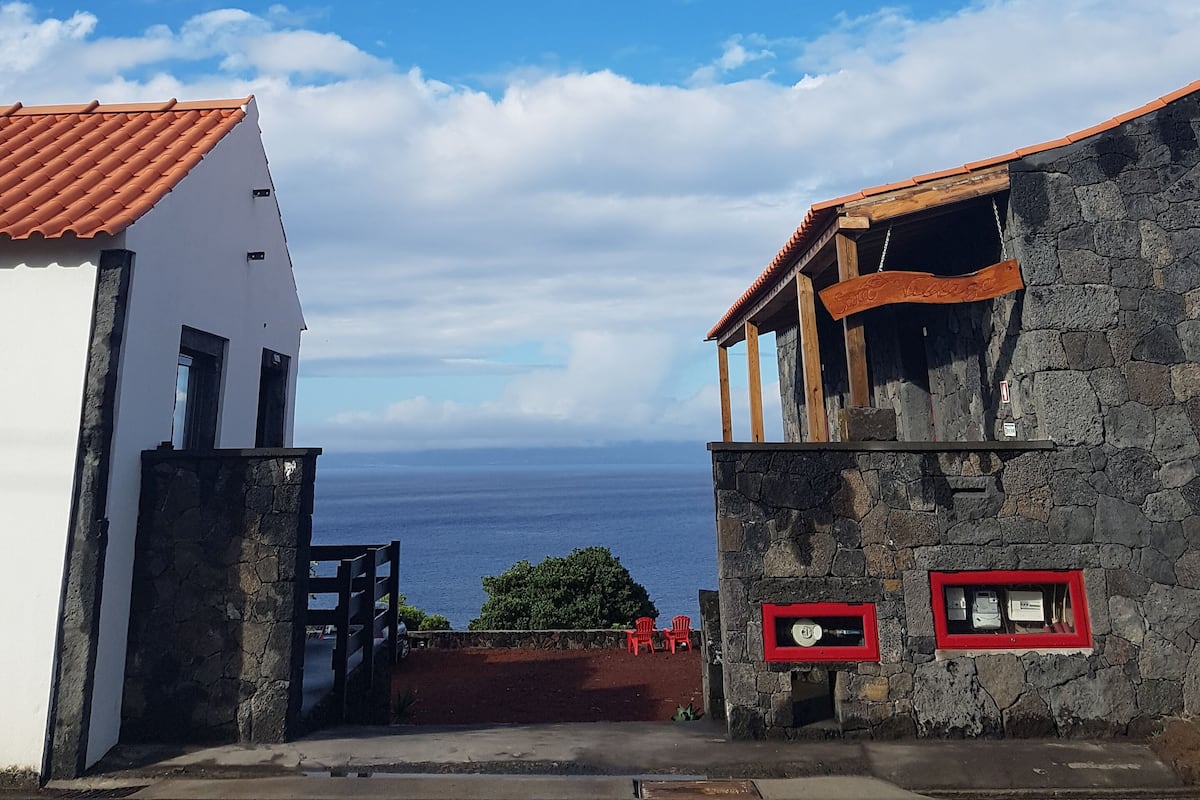 A coastal building with a stone facade and a red-tiled roof, featuring a wooden porch and a view of the ocean.