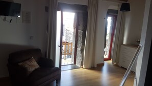 A living room with a sofa, a television, and a wooden ceiling. There are curtains on the windows, and a balcony is visible through the doorway. - Sara Village (Sao Roque do Pico)