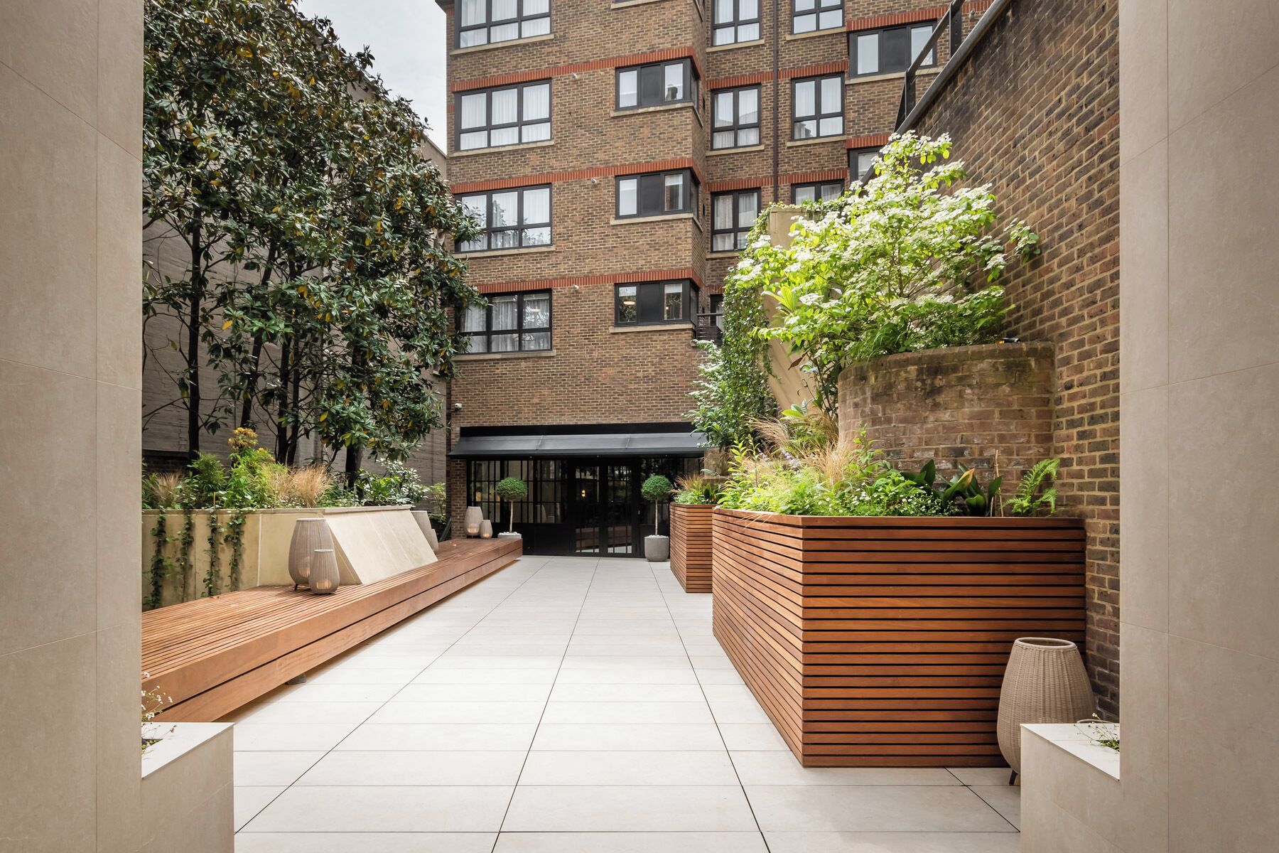 A modern urban courtyard with a brick building, wooden benches, and planters with greenery.