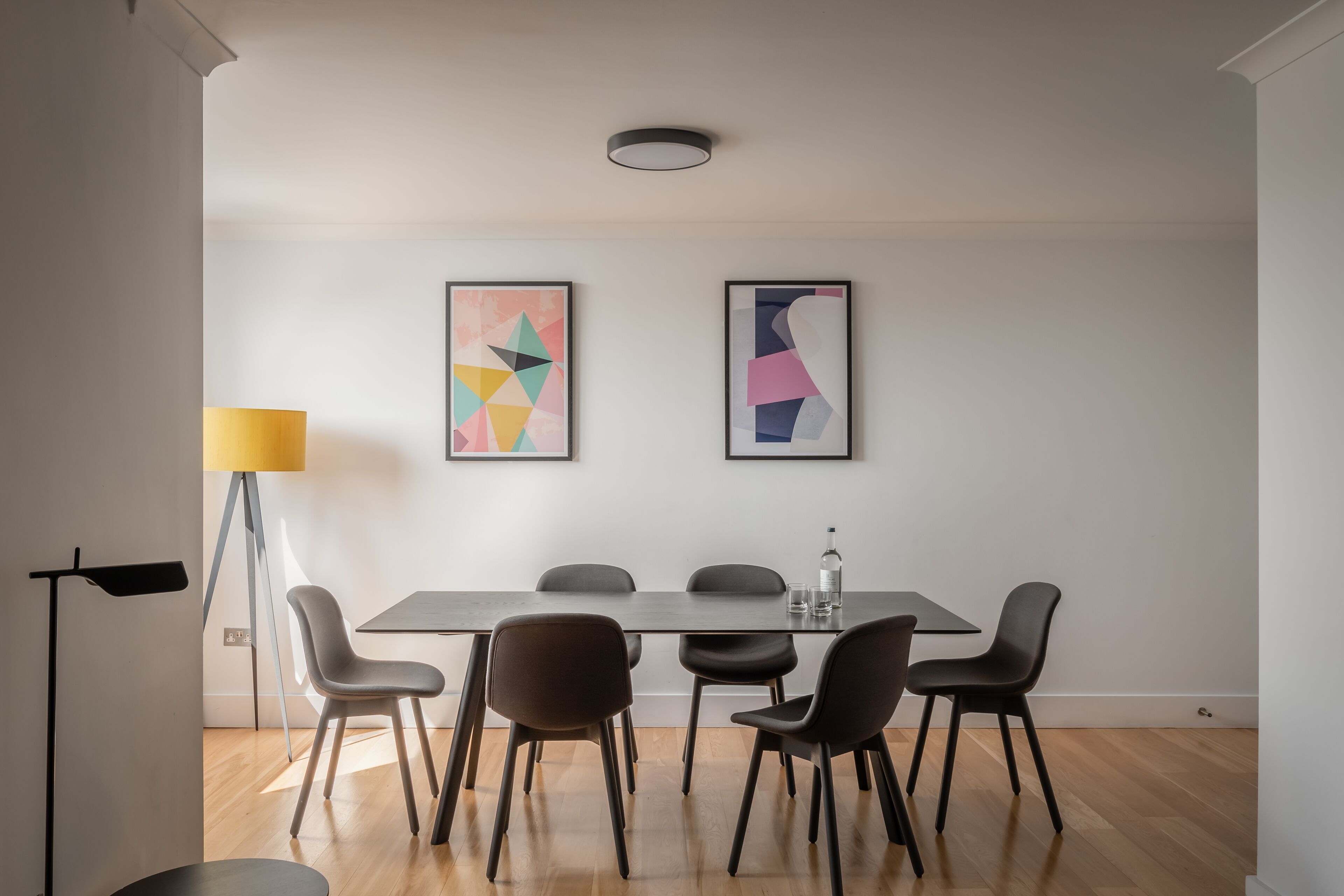 A modern dining area with a rectangular table, six chairs, two abstract paintings, a floor lamp, and a ceiling light fixture.