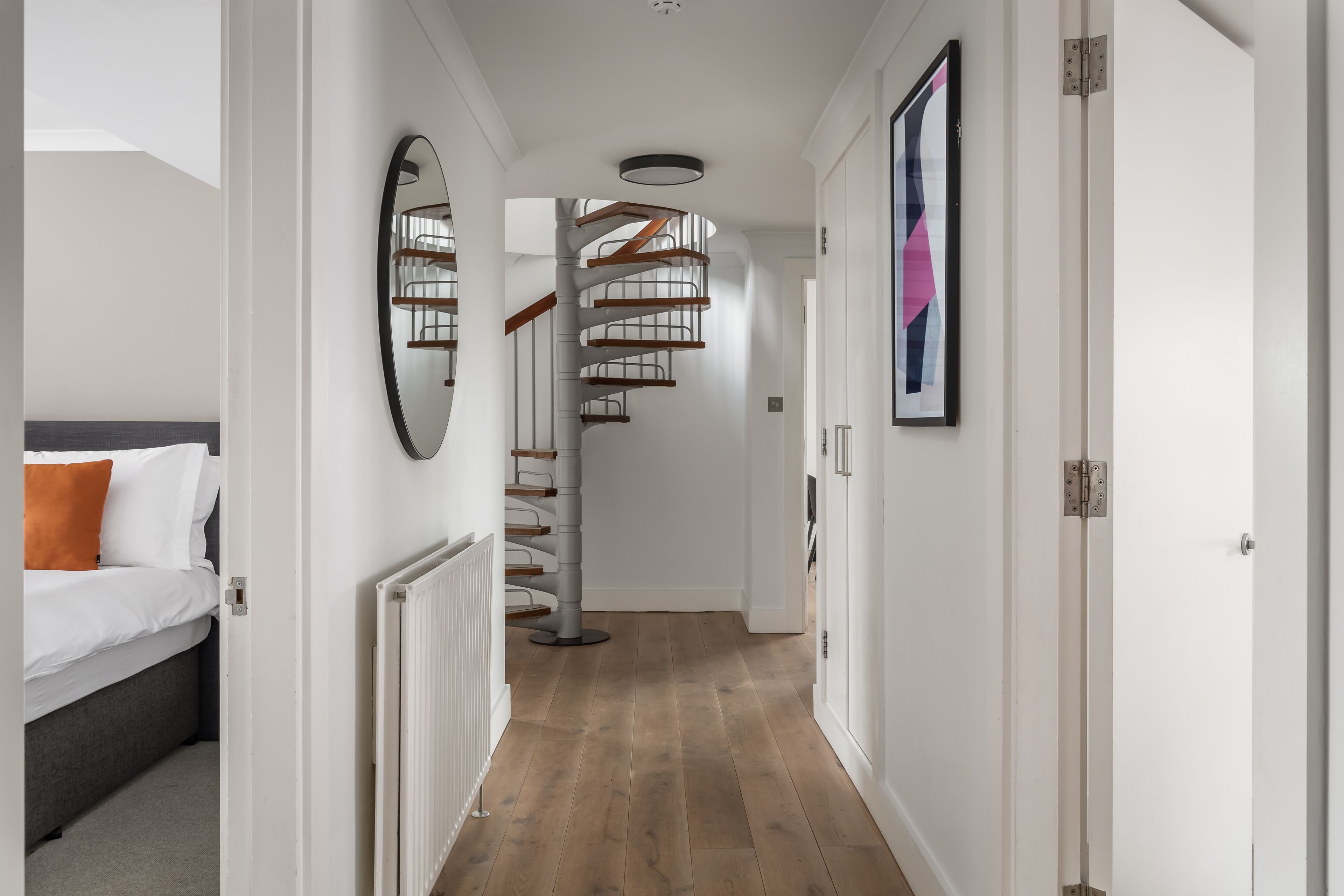 Un interior moderno con una escalera espiral, una cama con ropa de cama blanca y un cojín naranja, y un cuadro en la pared.