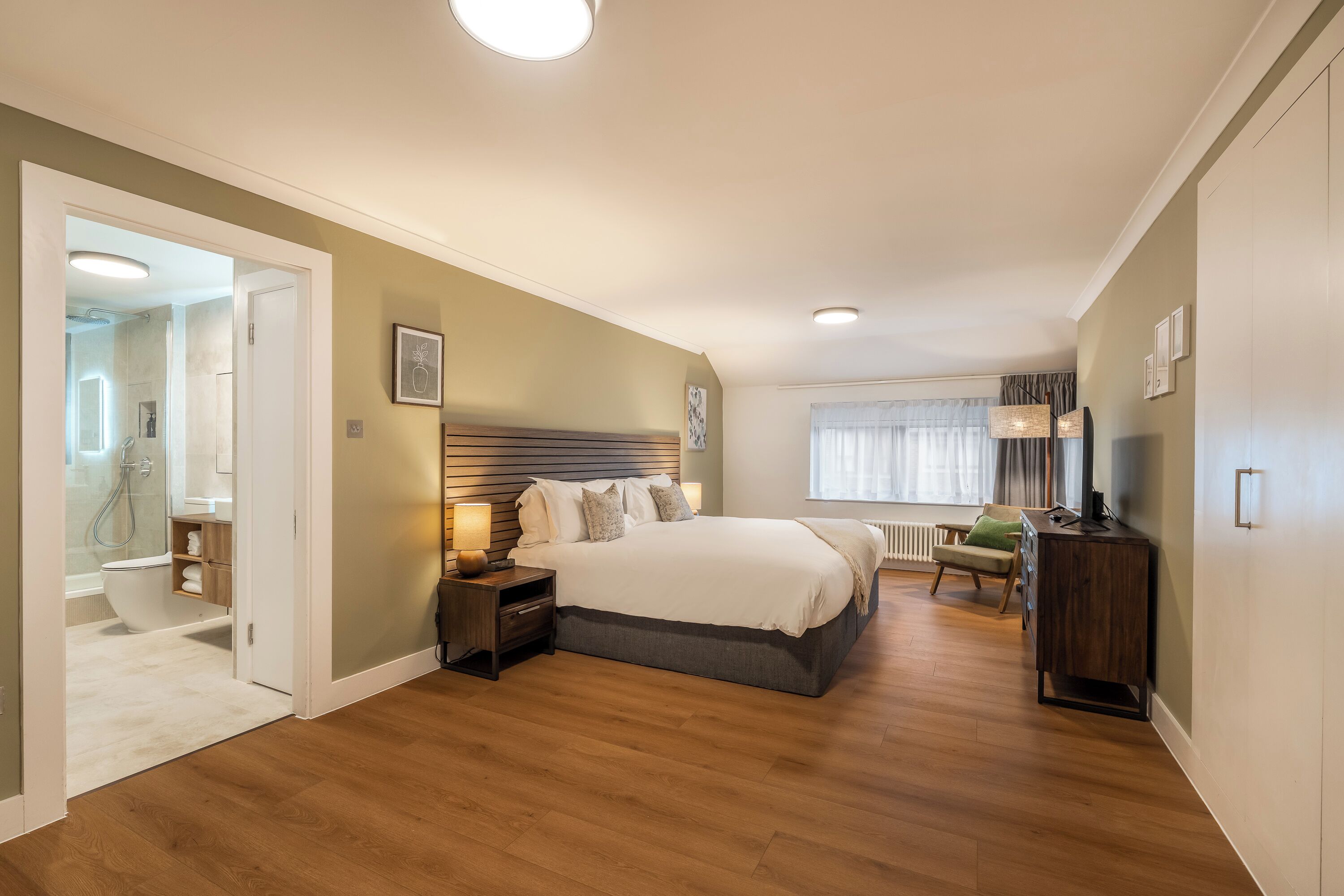 Un dormitorio con una cama grande, mesitas de noche, un aparador, una silla y una ventana con cortinas.