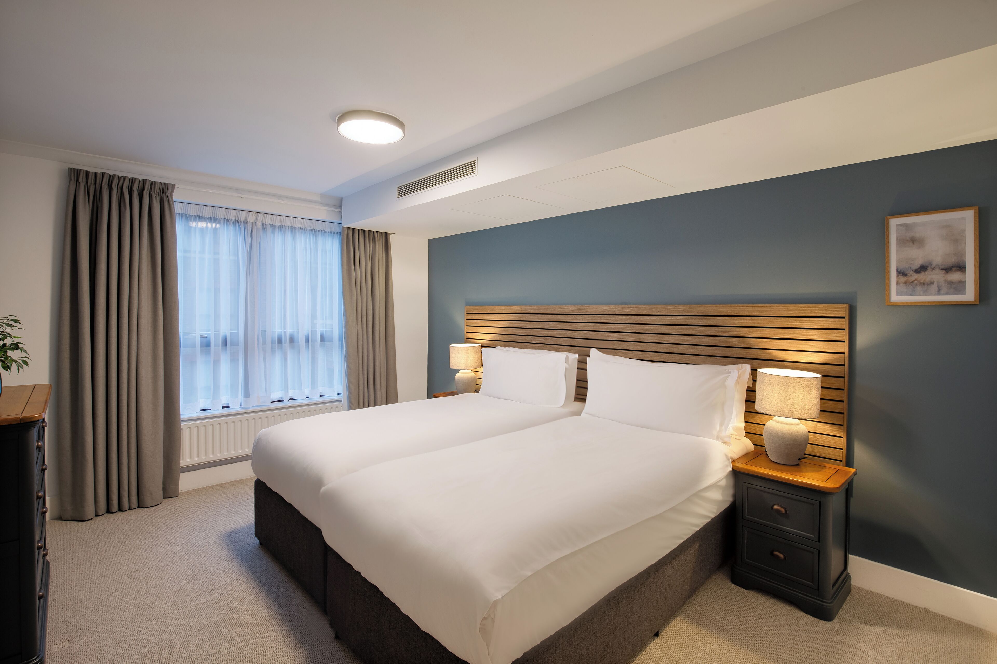 Une chambre d’hôtel avec un grand lit, des lampes de chevet, une tête de lit en bois et une vue sur l’extérieur à travers des rideaux transparents.
