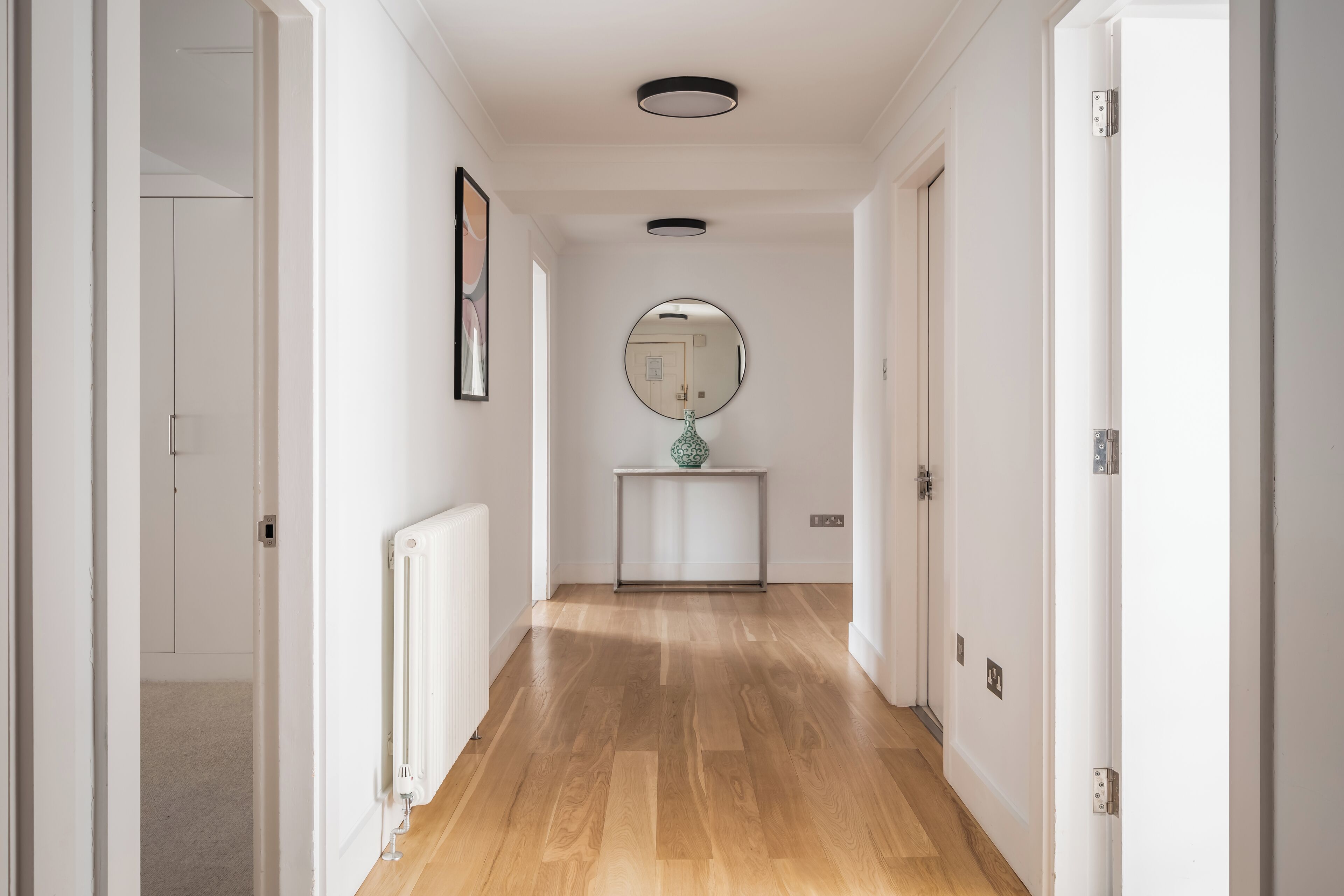 Een hal met houten vloer, witte muren, een radiator, een lijst met een afbeelding, een spiegel en een console tafel met een vaas.