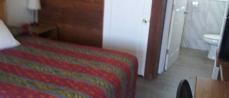 Un dormitorio con cabecera de madera, cama con colcha colorida, un escritorio con silla y un baño visible a través de una puerta abierta.
