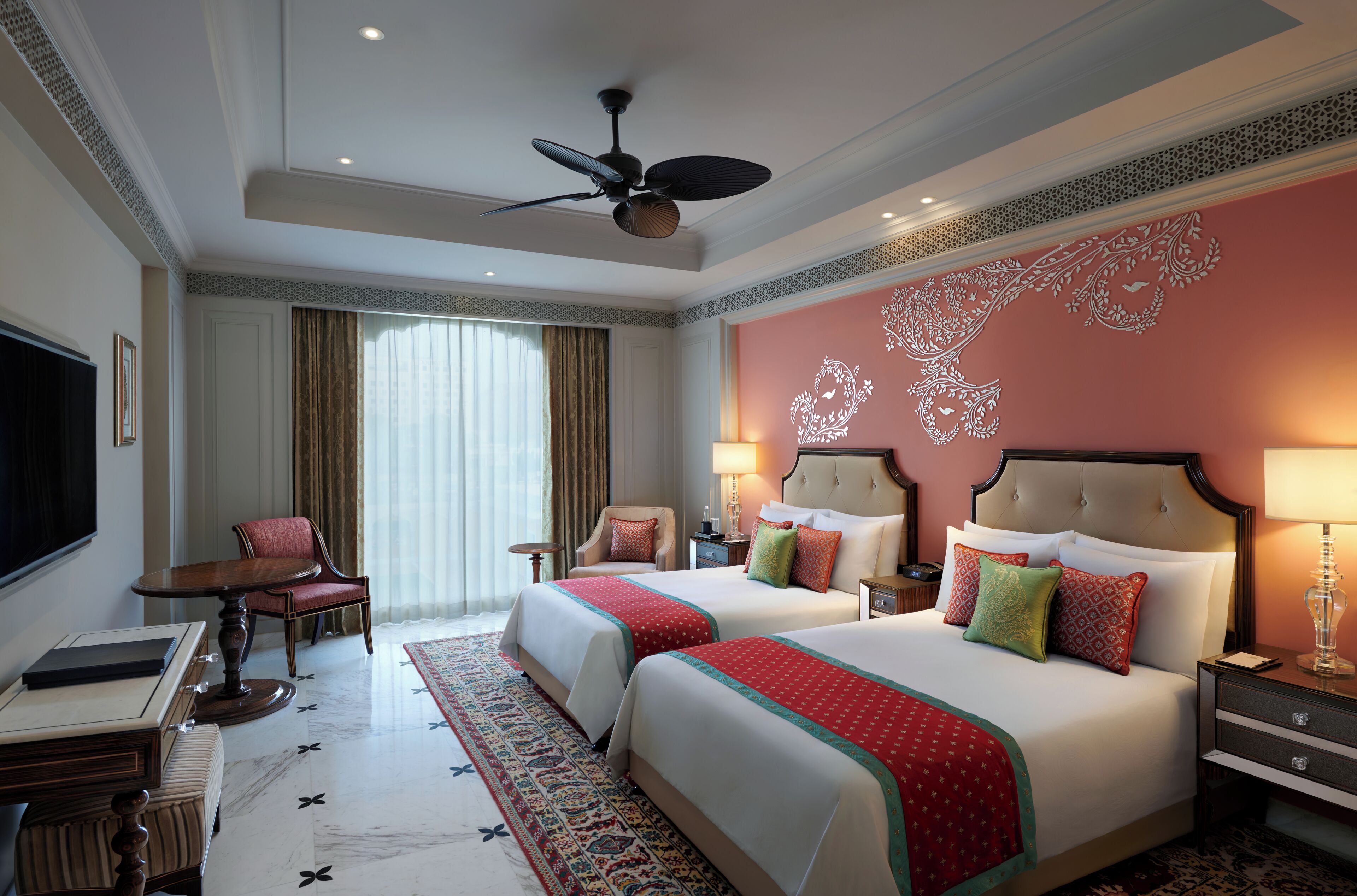 A hotel room with two beds, a desk, a chair, a TV, and a ceiling fan.