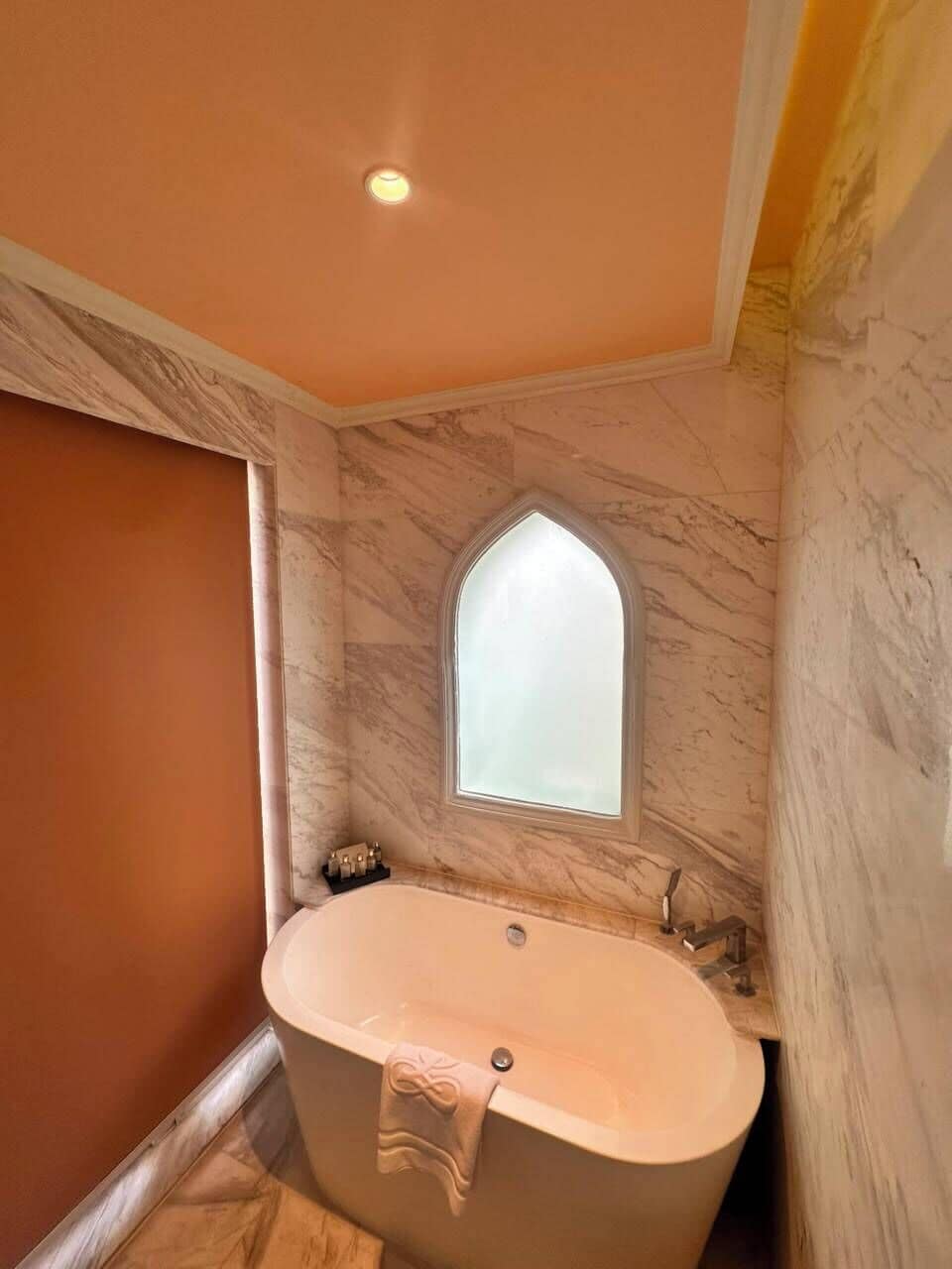 A bathroom with a freestanding bathtub, a small arched window, and marble walls.