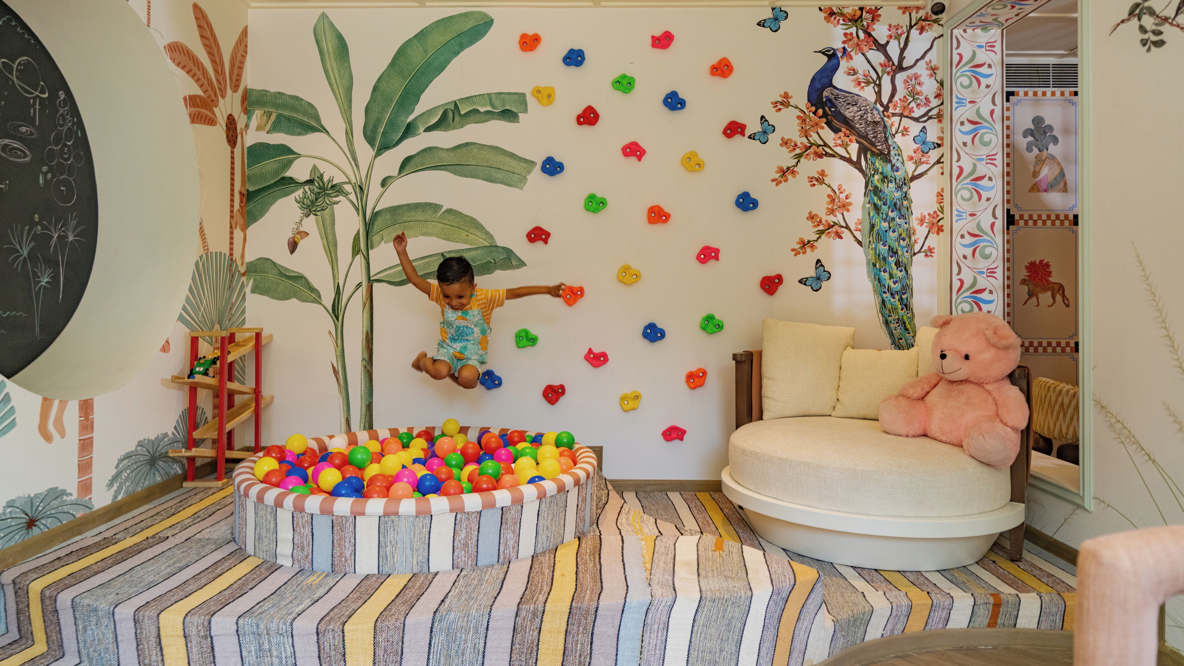 Un niño juega con pelotas de colores en una habitación con un área de descanso circular, una planta grande y adornos en la pared que incluyen un pavo real y mariposas.